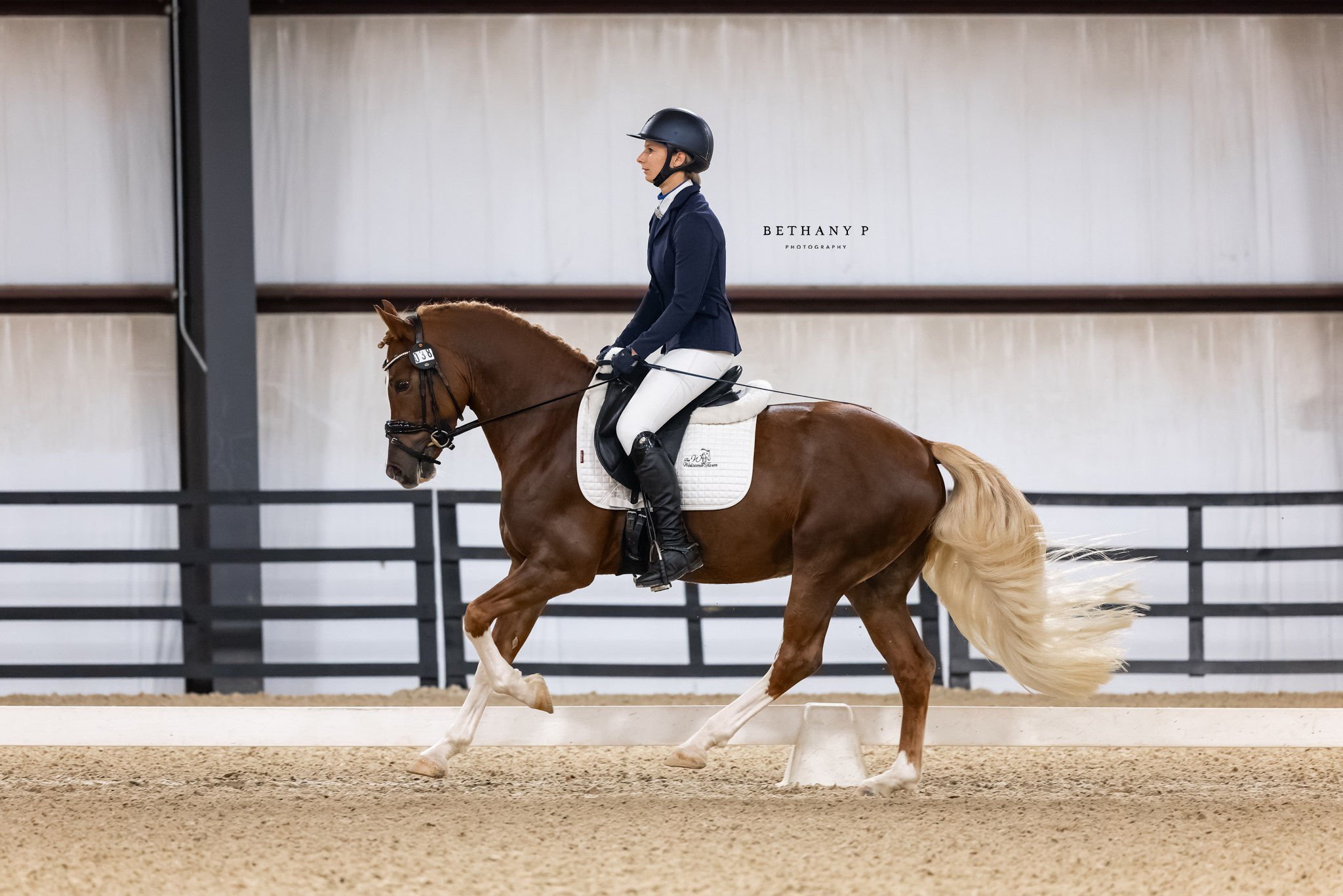 A female equestrian in riding gear riding a dressage horse with a white mane and tail in an indoor riding arena.