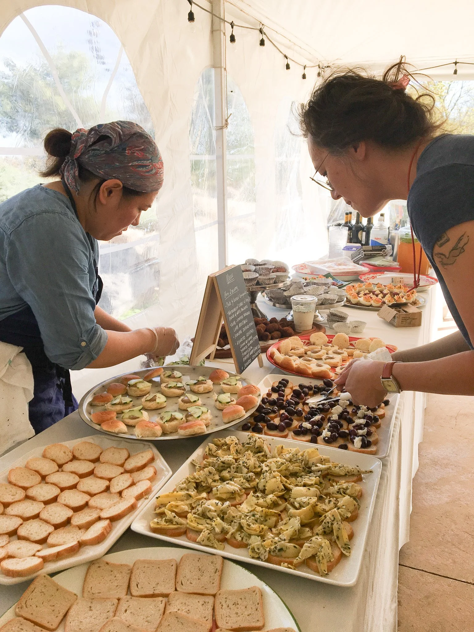 Harriet and Masae, of Leaf Kitchen in Iowa City, preparing and serving a variety of sandwiches and finger foods at a buffet table inside a tent with string lights.