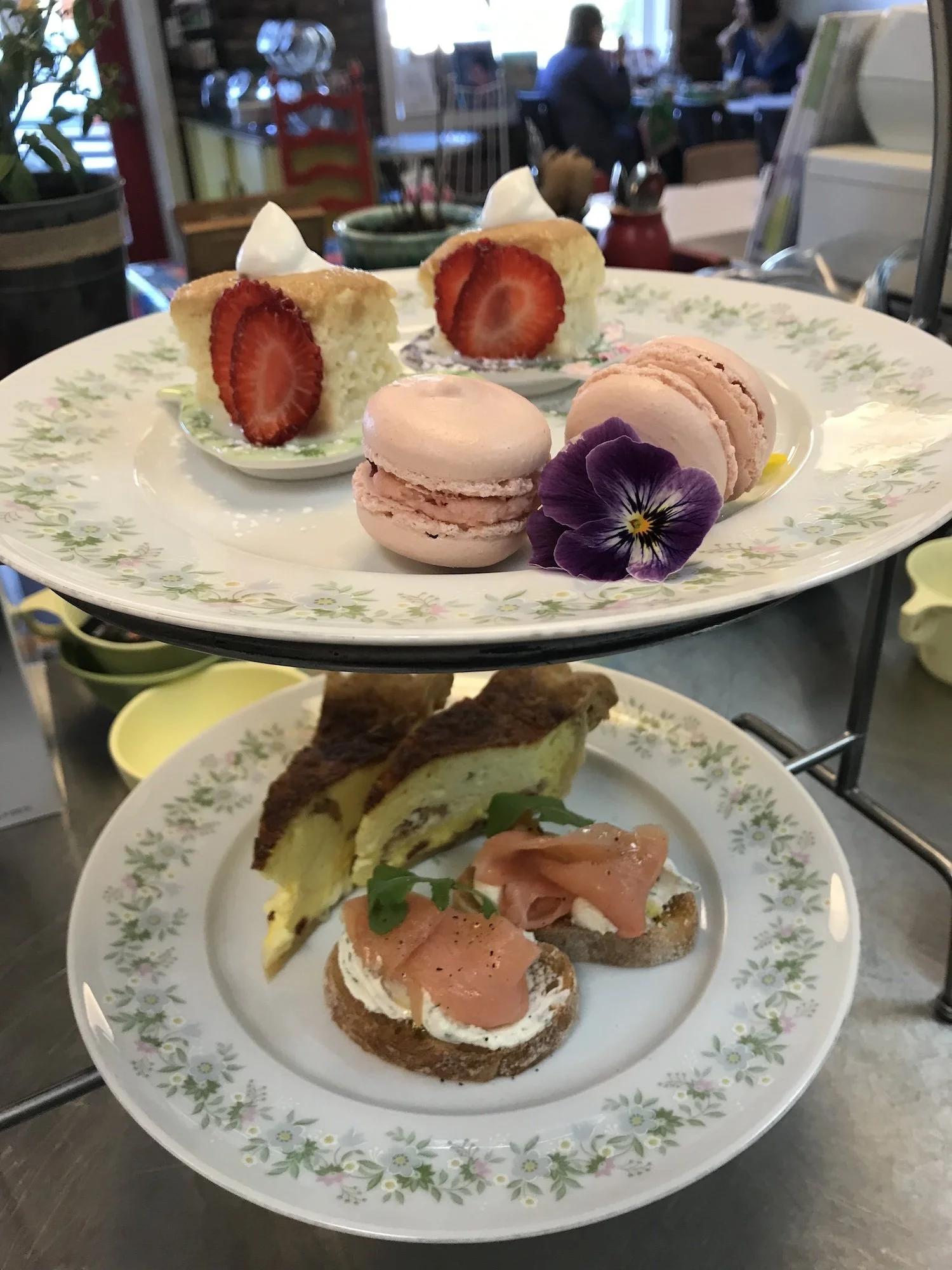 Two-tiered serving tray with assorted desserts at Leaf Kitchen's afternoon tea. Top tier with slices of strawberry shortcake, pink macarons, and a purple flower. Bottom tier with slices of savory quiche and toast with smoked salmon and cream cheese.