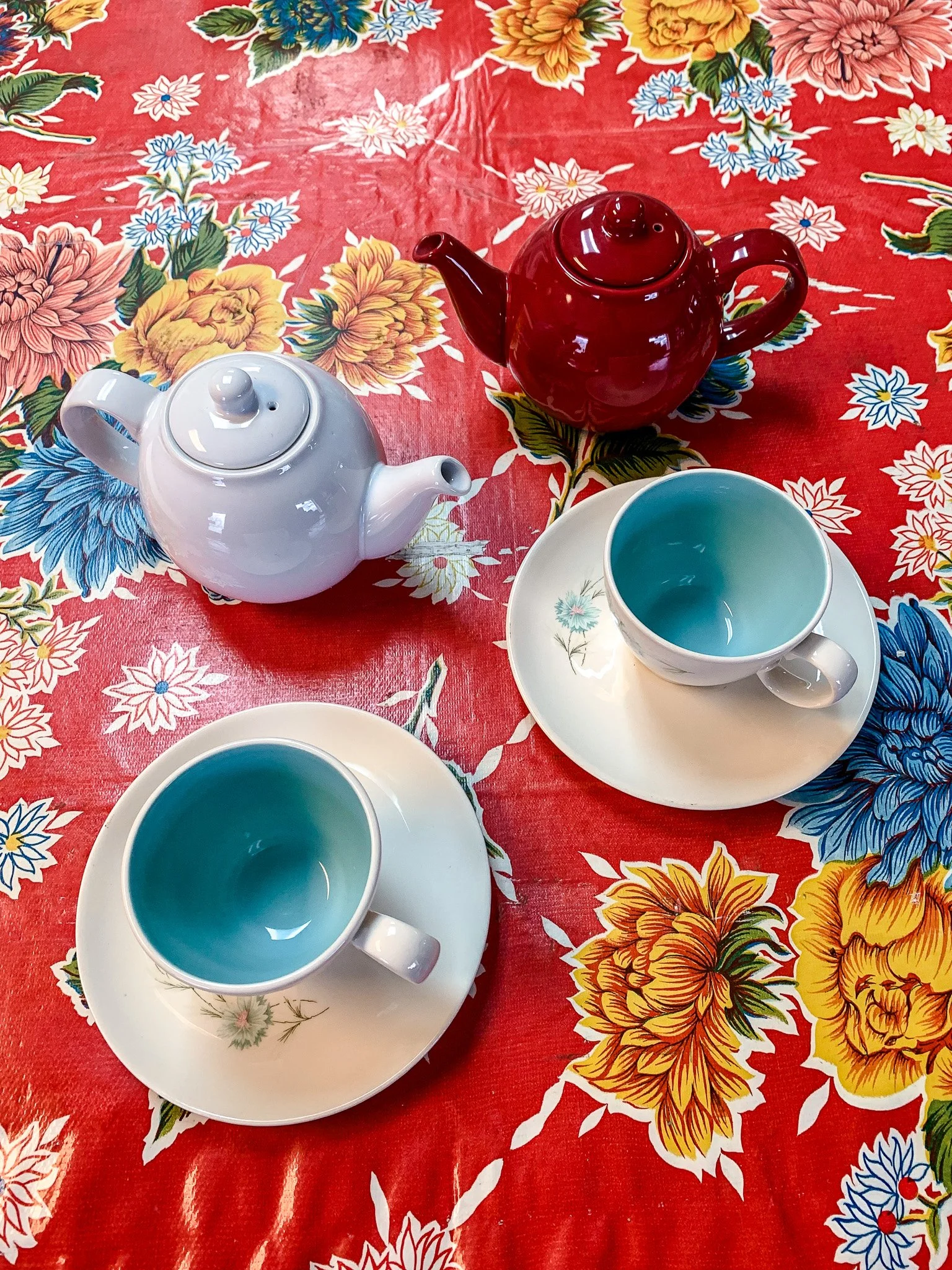 Two teapots, one red and one white, with matching cups and saucers on a vibrant, floral-patterned tablecloth.