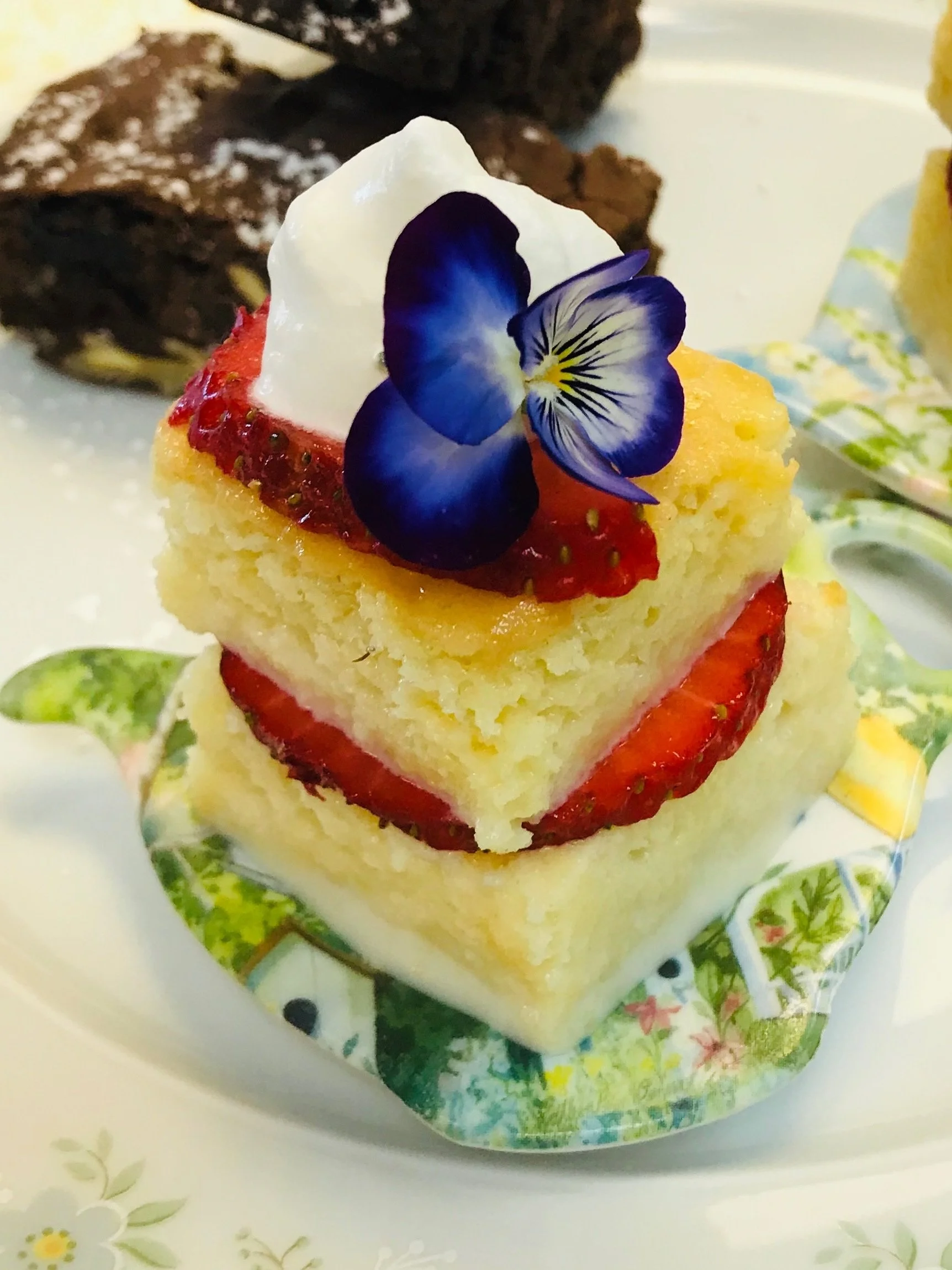 A small square cake with strawberries, topped with whipped cream and an edible flower, on a colorful dish, offered by Leaf Kitchen