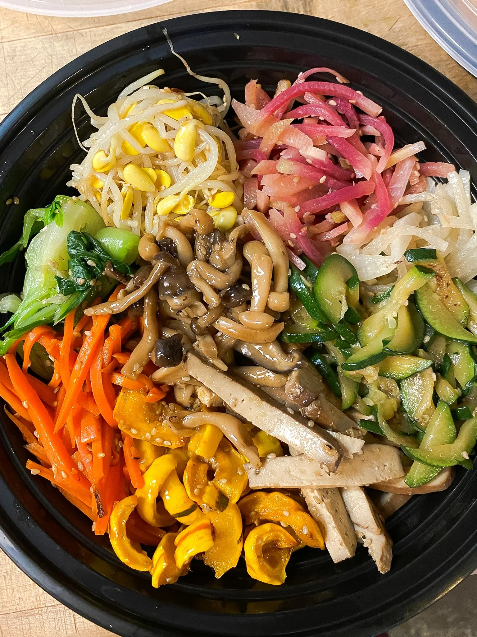 A take-home meal from Leaf Kitchen: A black salad container containing bibimbop: assorted fresh vegetables, including bean sprouts, pickled radish, shredded carrots, sliced zucchini, sliced cucumber, shredded cabbage, mushrooms, and sliced tofu.