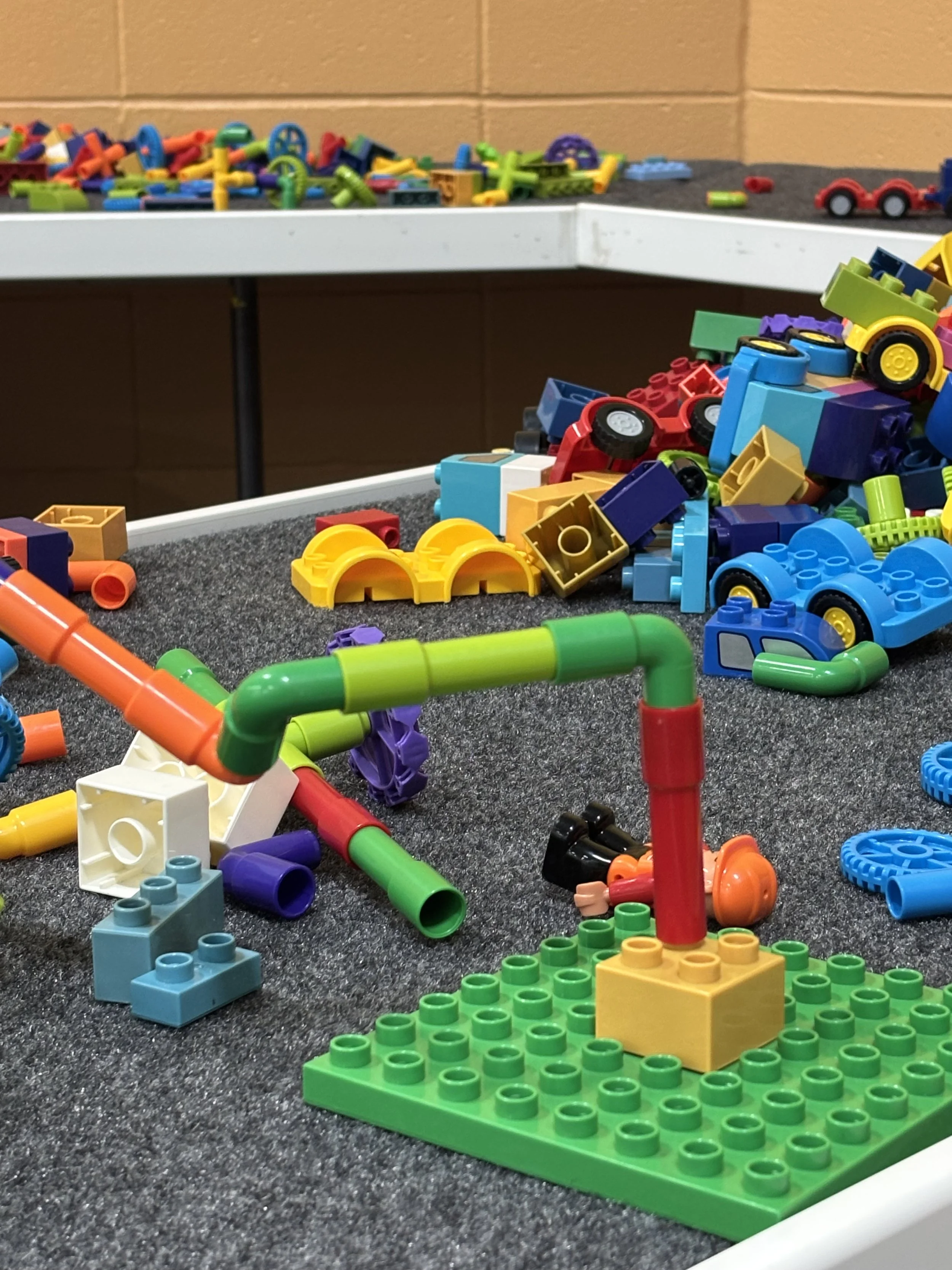 Colorful plastic building blocks and pipes scattered on a gray carpeted surface, with some built structures and loose pieces in the foreground and background.