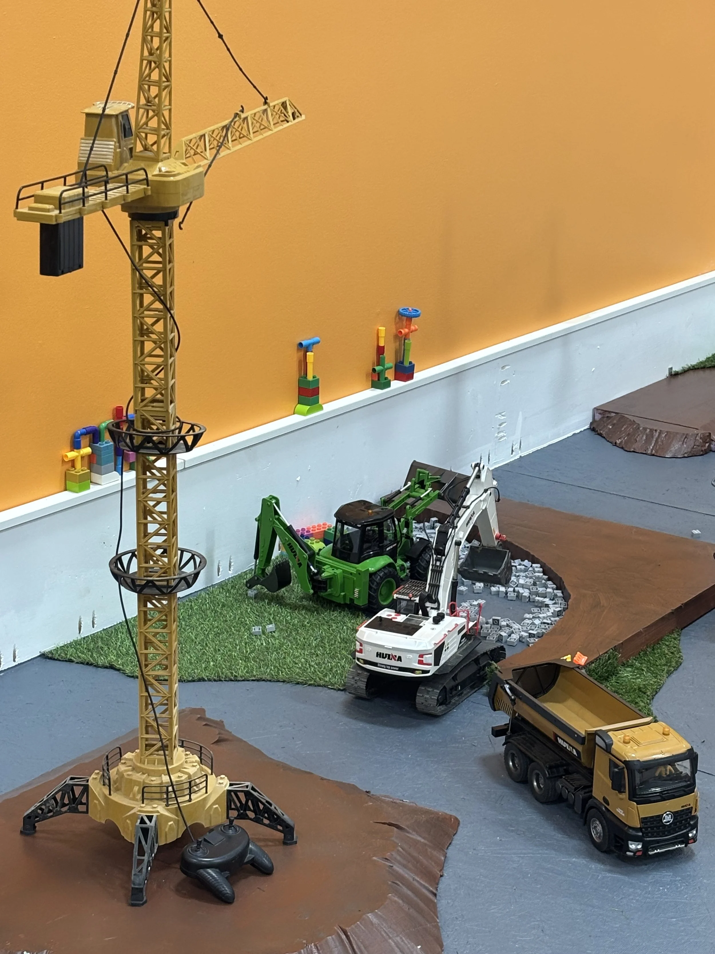 Toy construction scene with a tall yellow crane, toy vehicles including a green excavator, a white truck, and a yellow dump truck, on a surface with green grass and brown model terrain, and small toy building blocks against an orange wall.