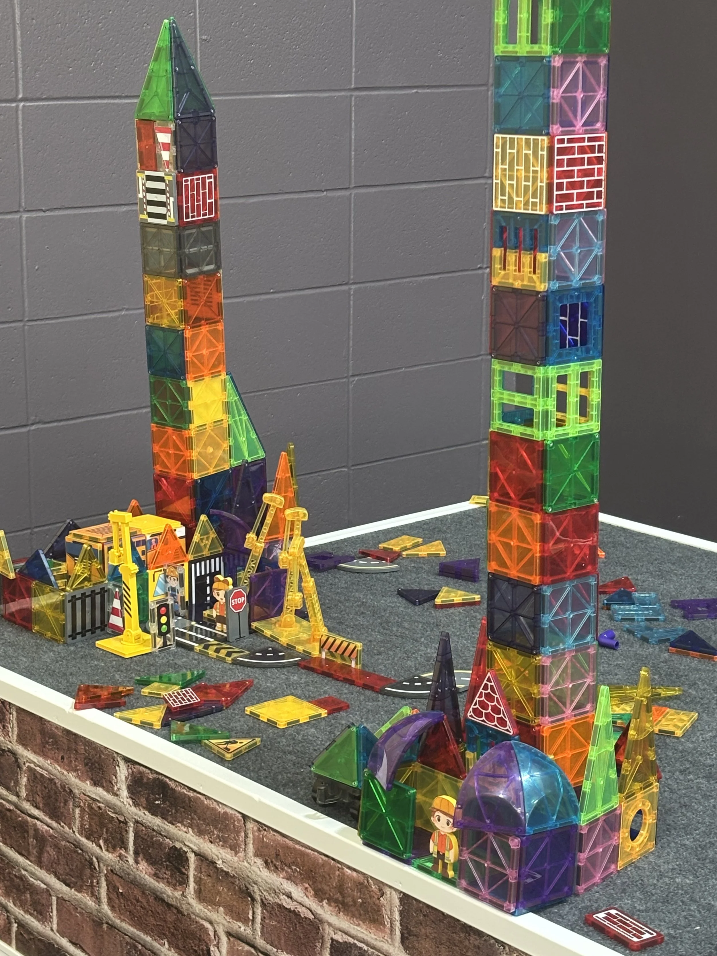 Colorful magnetic tiles arranged to create a cityscape and skyscrapers with various structures, vehicles, and miniature figures on a black platform against a gray brick wall.