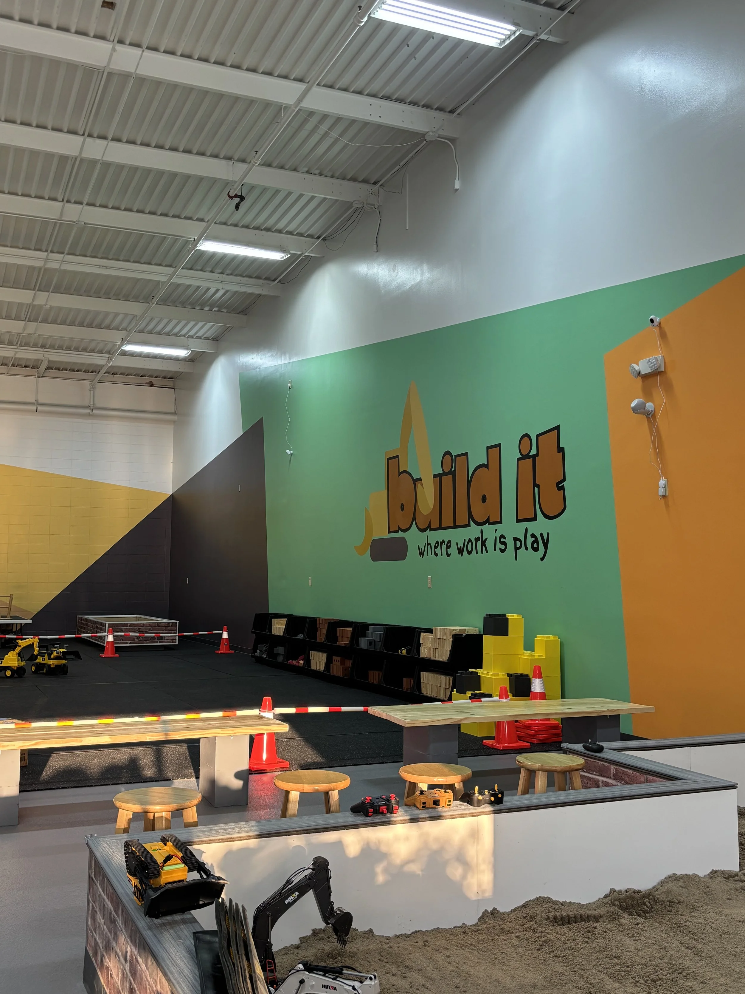 Indoor playground with colorful walls and construction toy area, sand box, small wooden stools, and construction equipment toys.