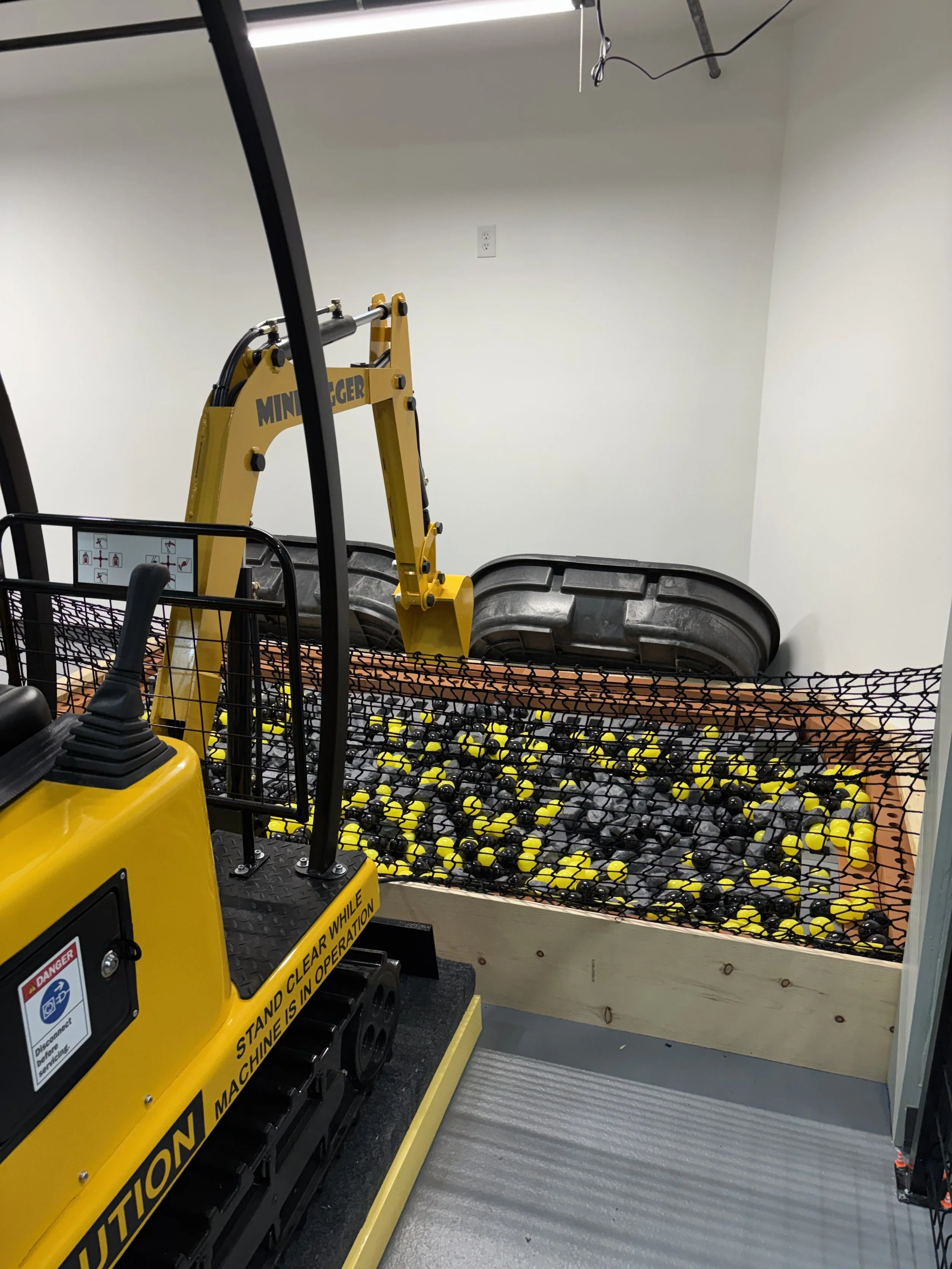 A small yellow mini excavator with a warning sign on its side, operating in an enclosed space with a black safety guard around the working area. The excavator is on a textured gray floor with a white wall in the background, which has an electrical outlet.