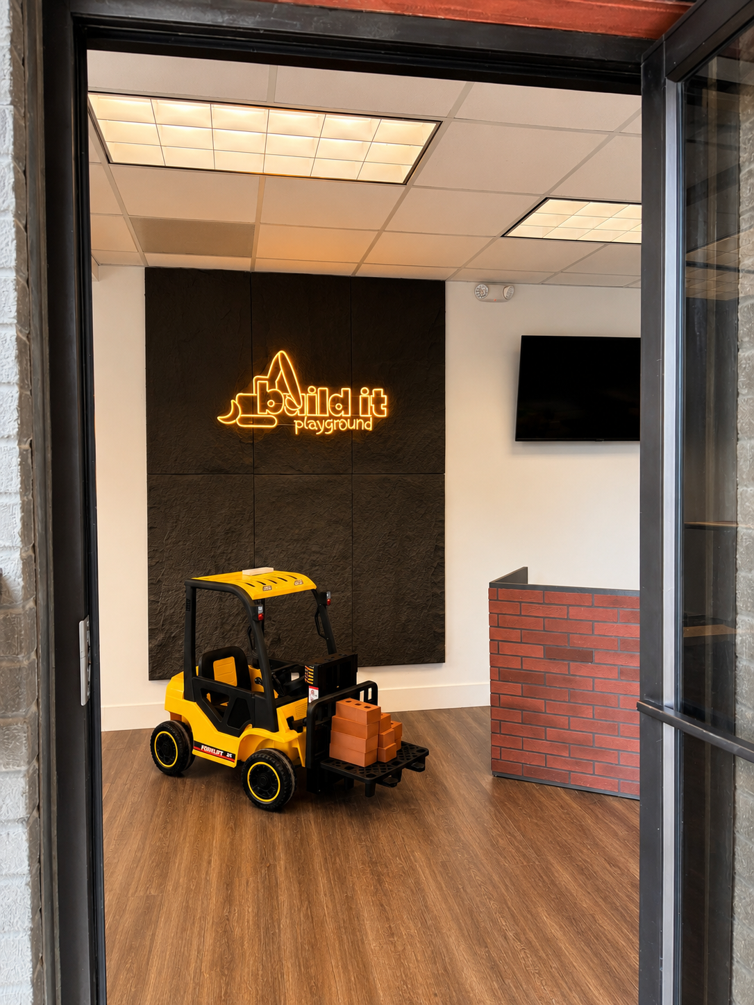 Toy forklift with bricks, neon sign reading 'Build it playground,' brick-patterned reception desk, wall-mounted TV, ceiling lights, and wood flooring.