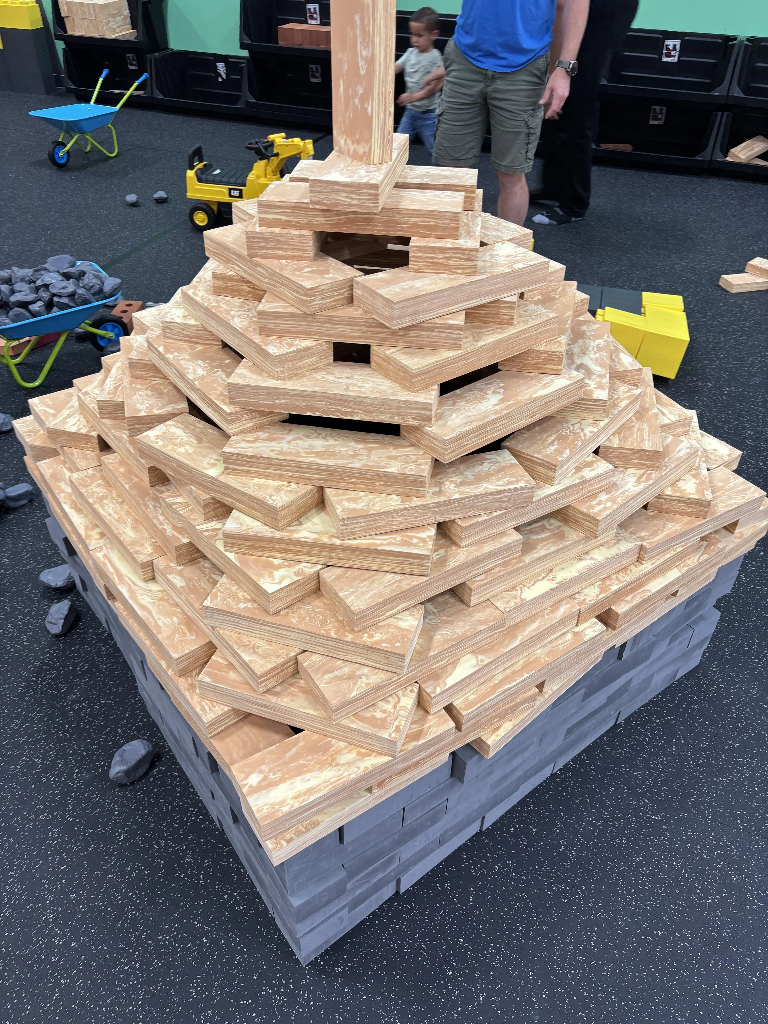 Stack of wooden building blocks in a pyramid shape, with some blocks missing. Toys and two children are in the background.