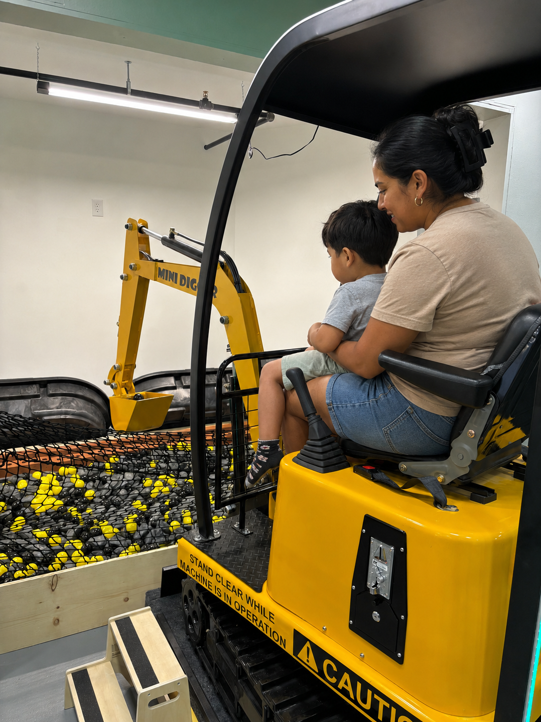 A woman and a young boy sit on a small yellow mini digger, with the woman guiding the boy as he plays with the controls. The mini digger has a black metal arm and a yellow bucket, and there is a warning sign on its side.