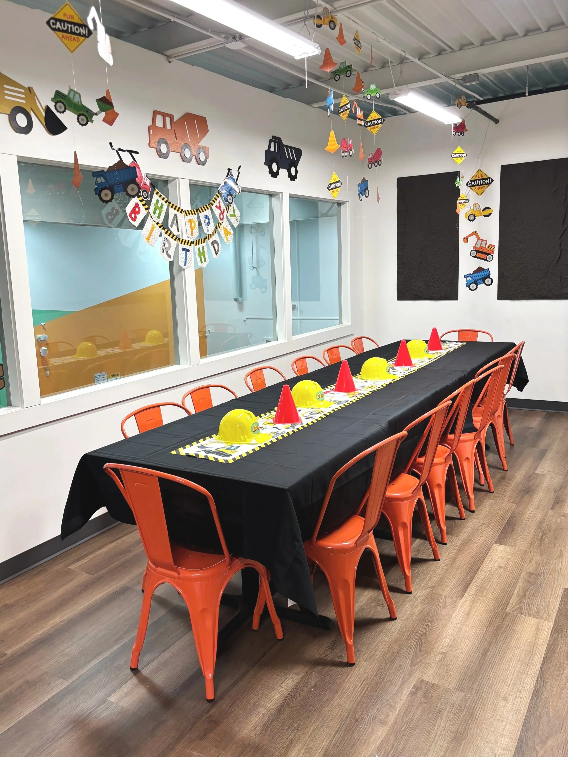 Party setup with a long table covered in a black tablecloth, orange chairs, yellow construction helmets, red napkins, and a
