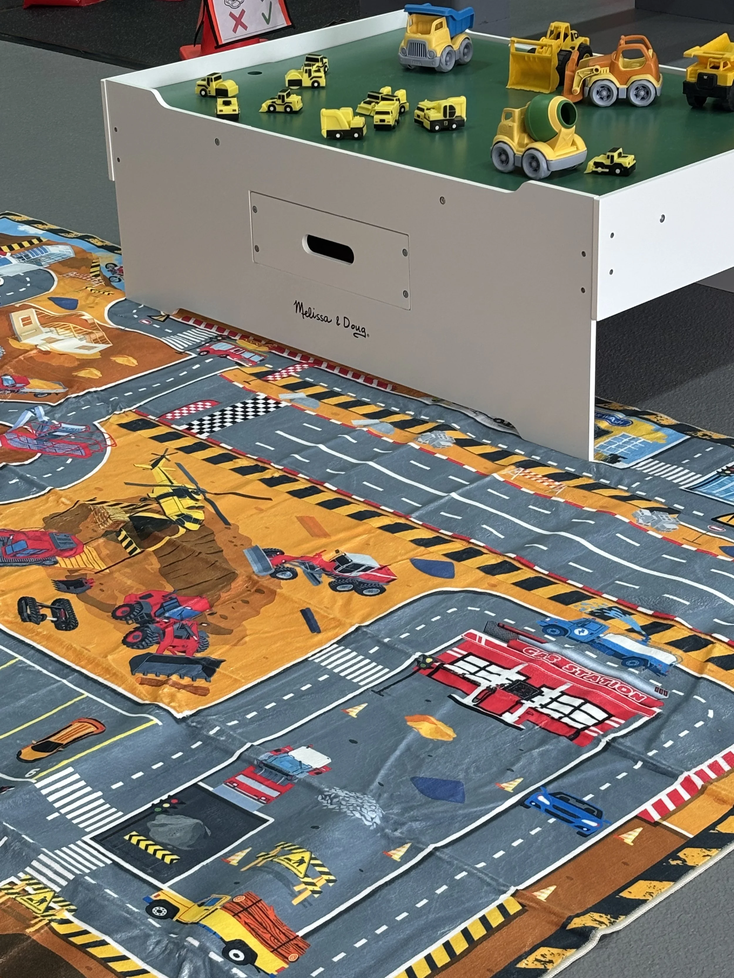 A play mat with roads, traffic signs, and construction zones, decorated with toy vehicles like fire trucks, construction equipment, and other construction-themed toys, situated beneath a white storage bin with small toy vehicles on top.