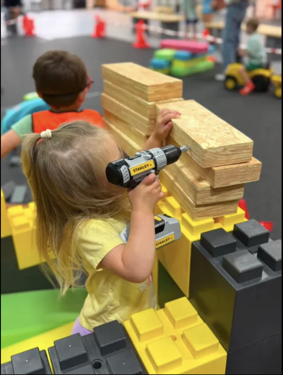Children playing with large building blocks and toy tools in a play area.