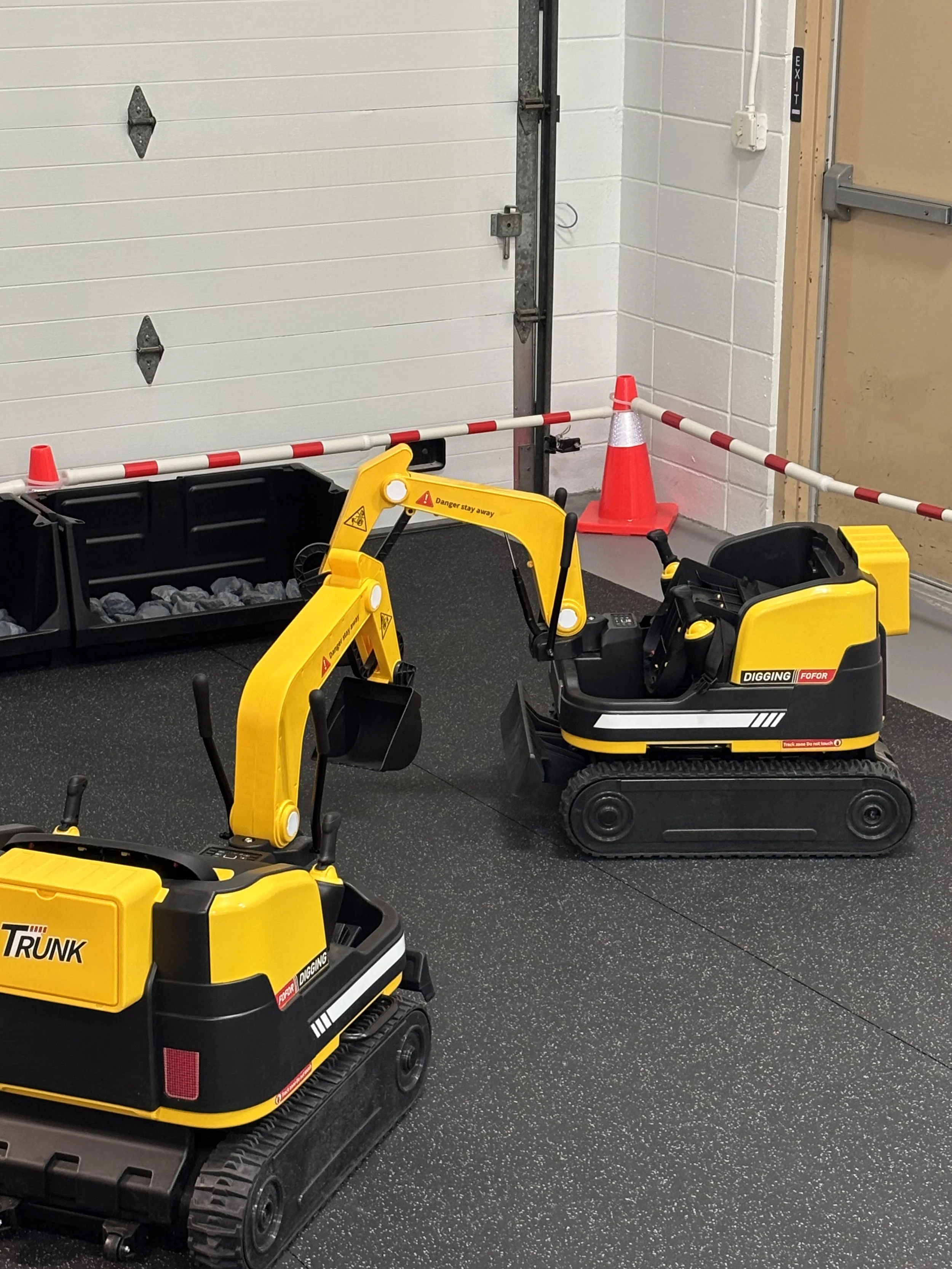 Children's toy construction equipment, including yellow excavator and black dump truck, in a play area behind safety cones and barrier at an indoor facility with white cinderblock walls.