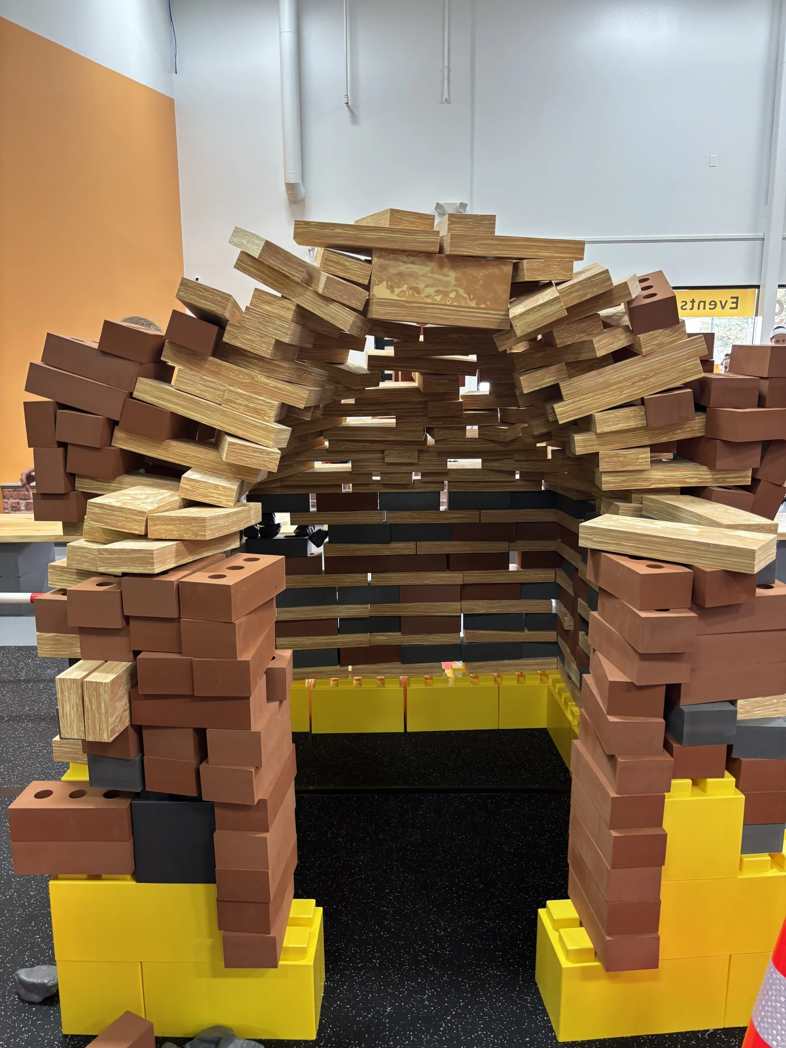 Large LEGO sculpture resembling a person wearing a hat, made with bricks in various shades of brown, black, yellow, and gray, displayed in an indoor setting.