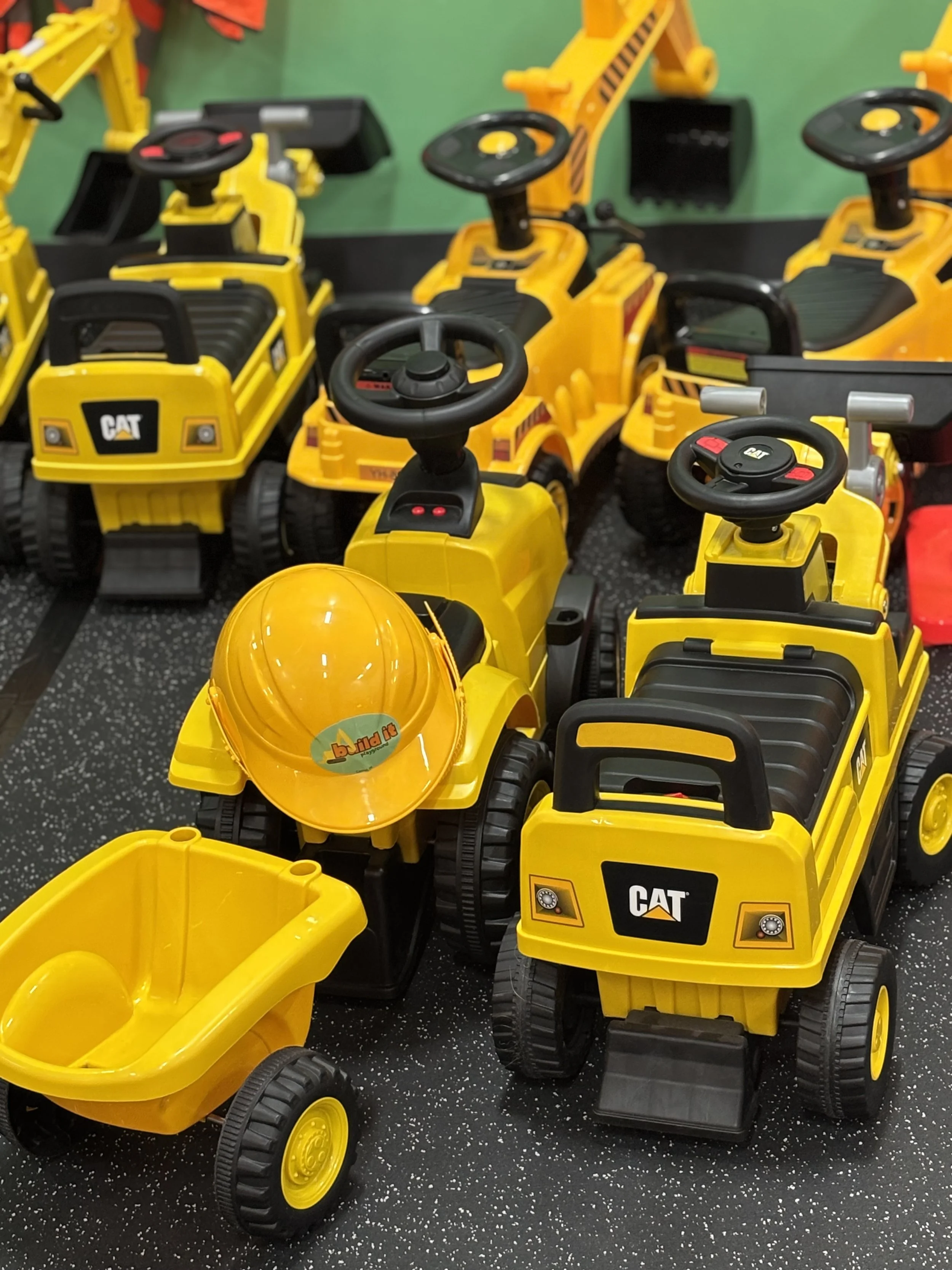 Multiple yellow toy construction vehicles, including ride-on trucks and a dump truck, arranged on a black speckled floor, with a green wall background.