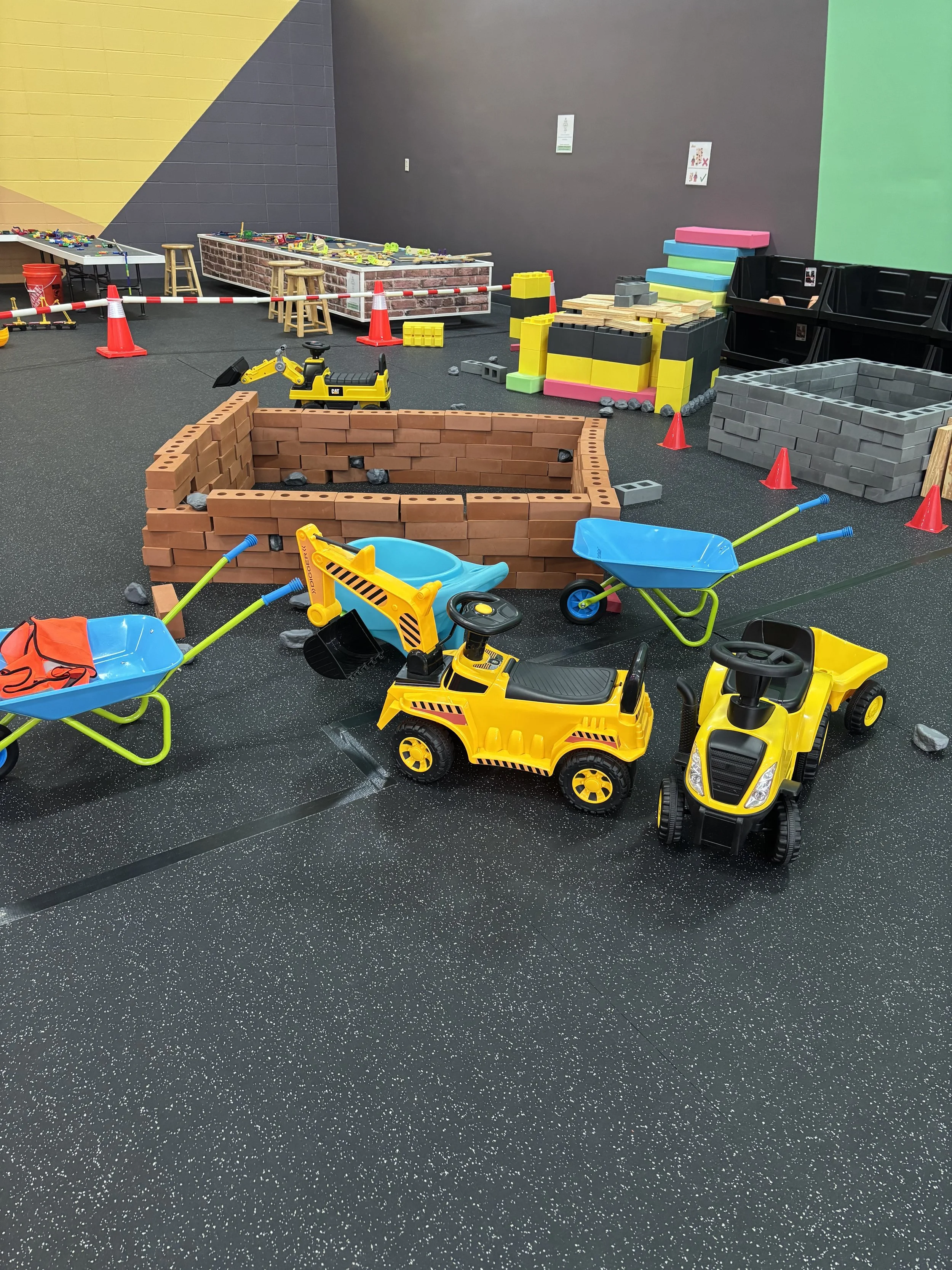 Children's indoor play area with toy construction vehicles, building blocks, and colorful foam mats, sectioned off with orange cones.