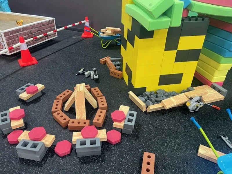 Colorful indoor play area with LEGO-like blocks, a rock pile, construction cones, and tools.