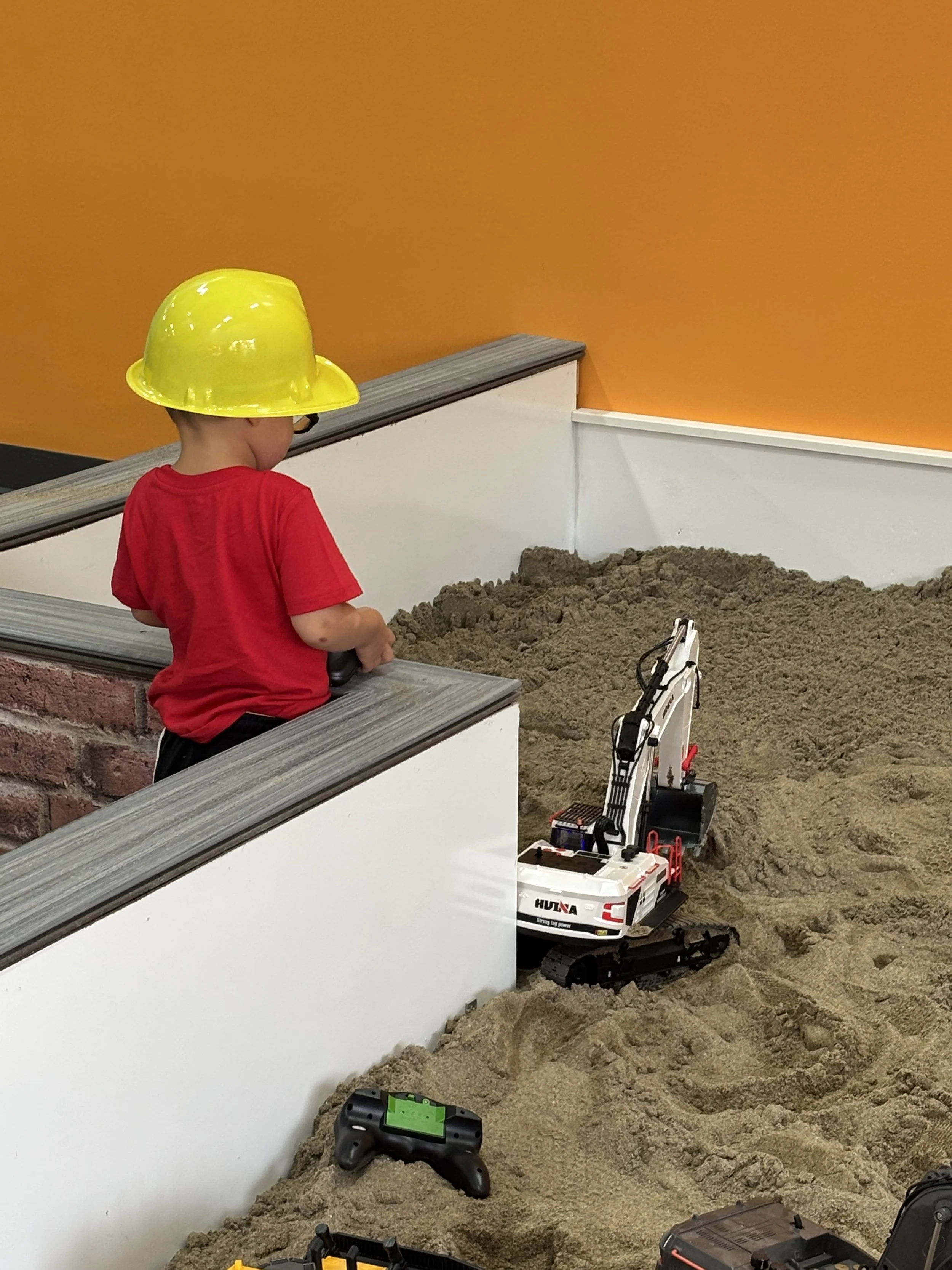 Child wearing a yellow construction helmet and a red shirt playing with a remote-controlled toy excavator in a sandbox with construction-themed toys and tools.