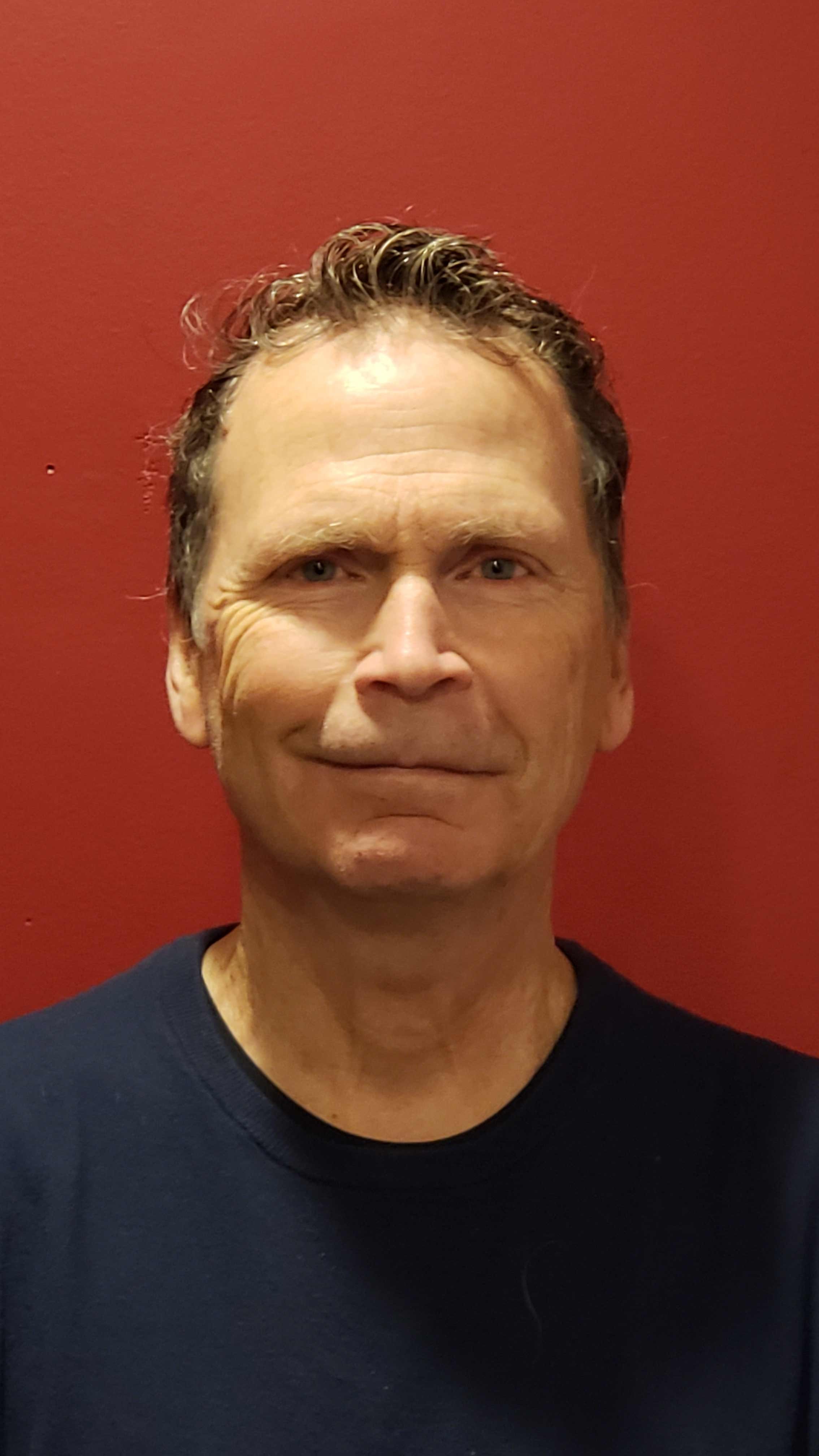A middle-aged man with short, wavy brown hair and blue eyes, wearing a black shirt, standing against a red background.