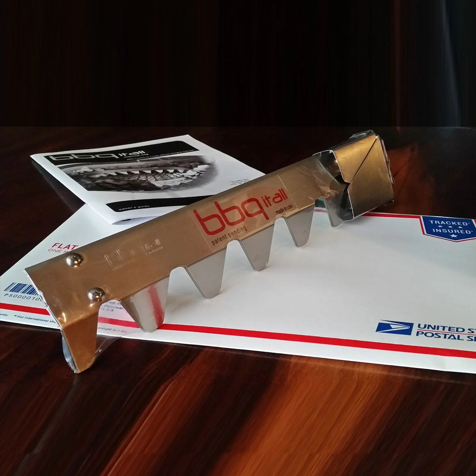 A metal grill with 'bbq it all' branding resting on USPS mail envelope.