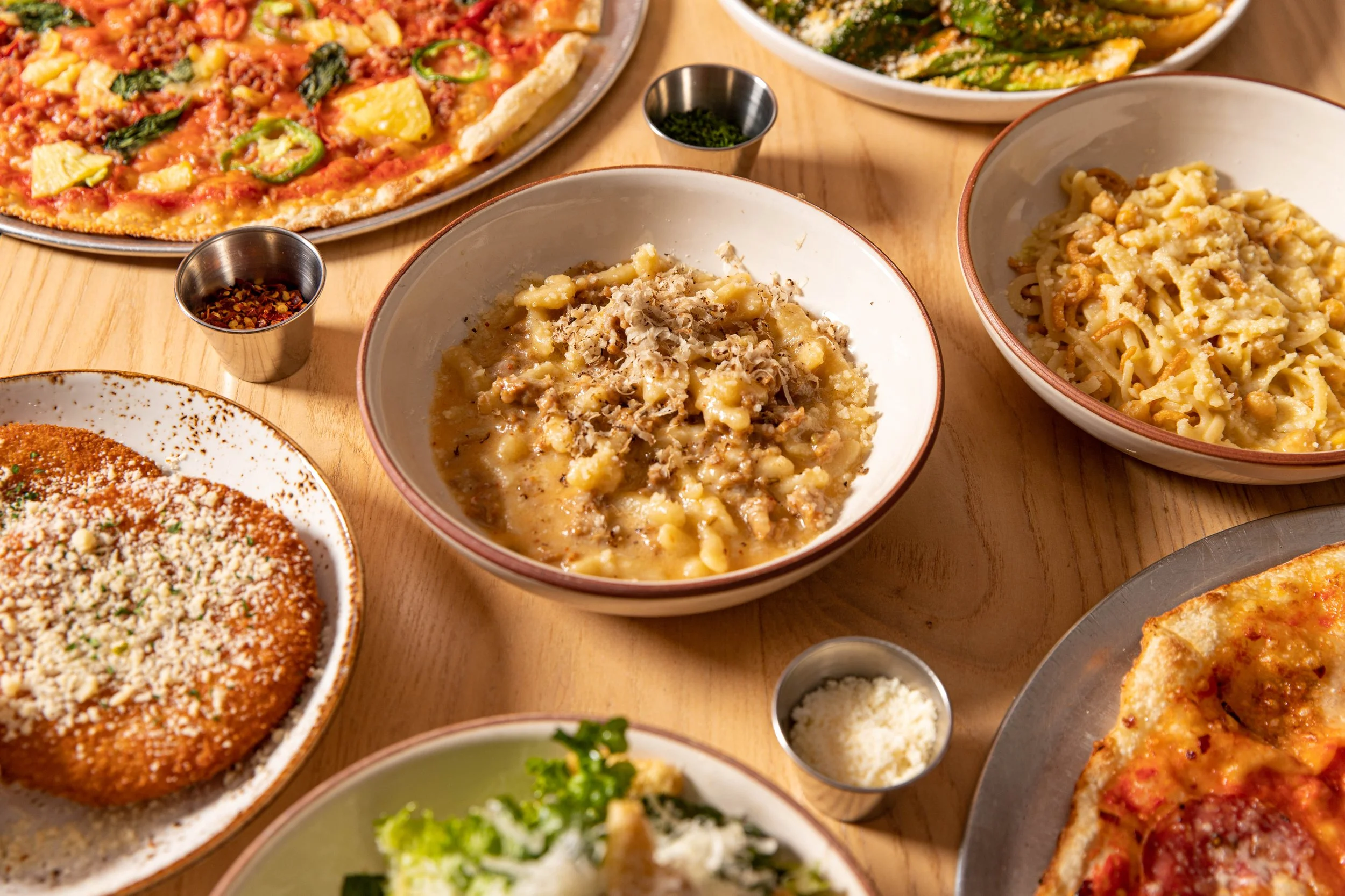Various dishes of Italian food on a wooden table, including a pizza, a bowl of ravioli, a bowl of gnocchi, a plate of chicken parmesan, and a slice of pepperoni pizza with cheese.