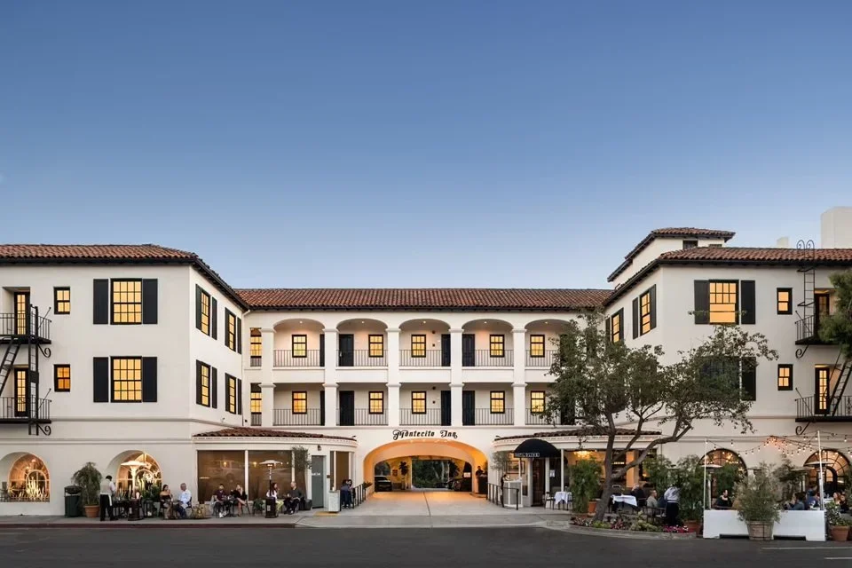 The Montecito Inn, a white three-story hotel with black window shutters and a red tile roof, features an arched entrance and outdoor seating areas on both sides, with people dining and walking under string lights and tree shade.