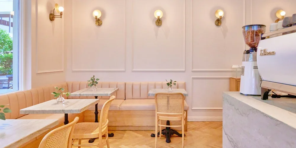 Interior of a café with cream-colored walls, gold wall sconces, marble tables, natural wood and cane chairs, beige bench seating, small plants in glass vases, and a coffee station with a grinder and espresso machine.