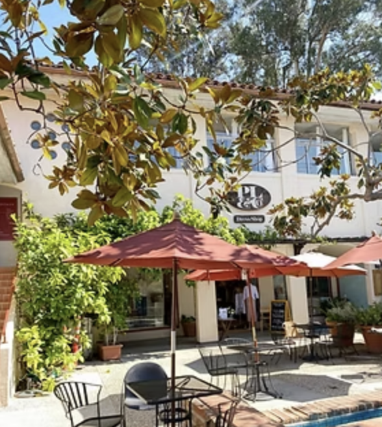 Outdoor patio with tables, chairs, and umbrellas in front of a cafe or restaurant, surrounded by greenery and trees.