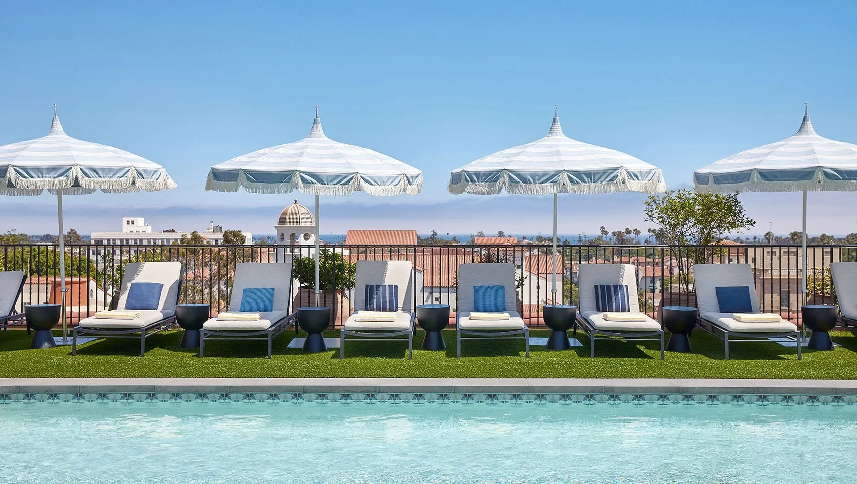 Poolside patio with white lounge chairs, navy pillows, black side tables, and white parasols with fringe, overlooking a cityscape with rooftops and a water view in the distance under a clear blue sky.