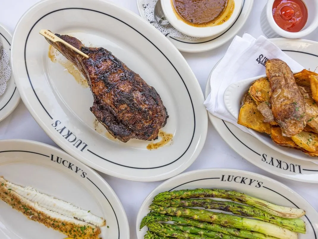 Meal with grilled lamb chop, grilled asparagus, mashed sweet potatoes, and side dishes of sauces and desserts on white plates with Lucky's logo.