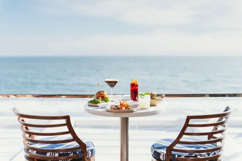 A round table with two chairs on a balcony overlooking the ocean, set with various meals and drinks.