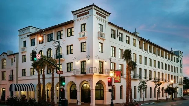 A white multi-story building with arched entrances, palm trees, and decorative balconies, illuminated during sunset.