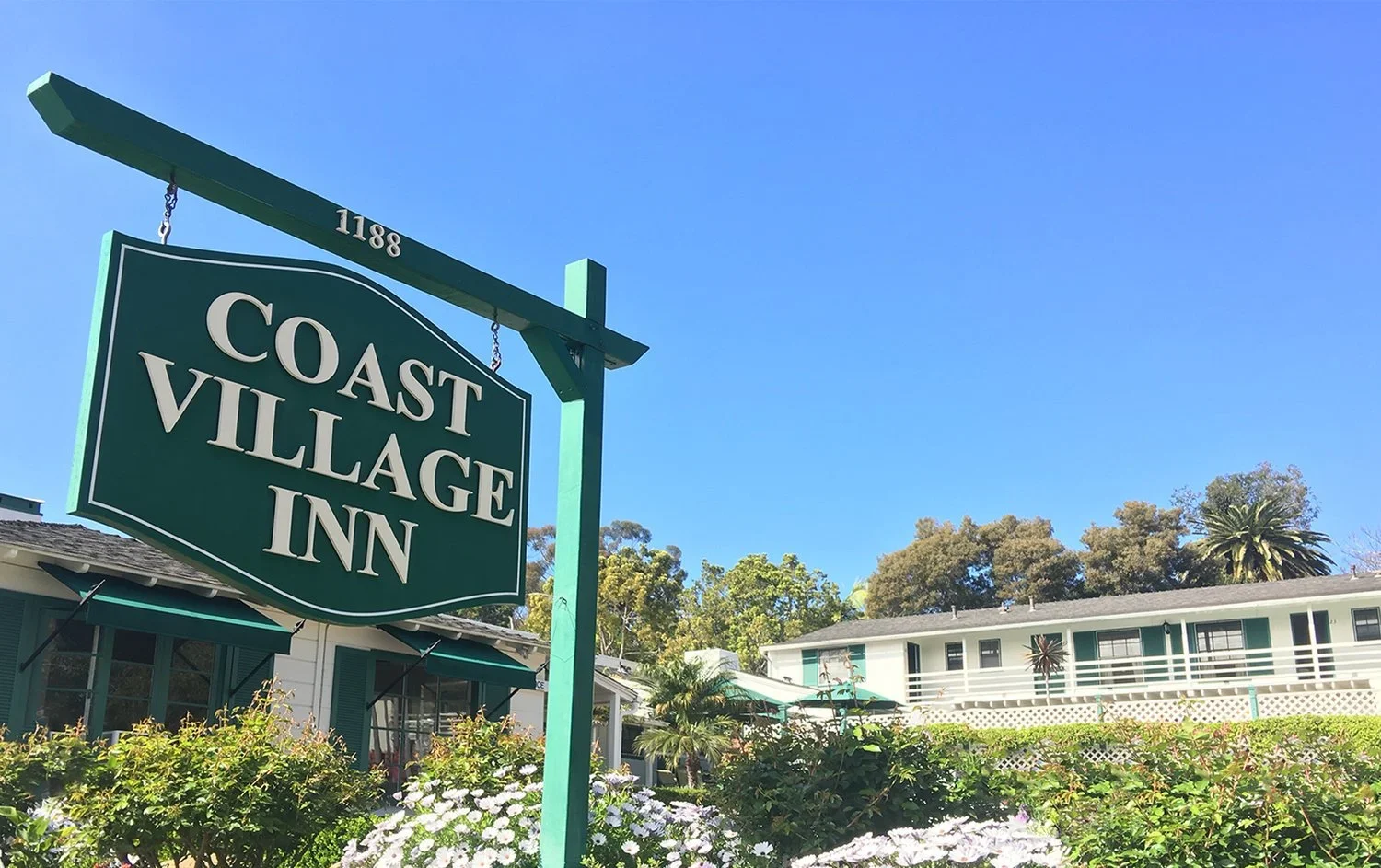 Green sign for Coast Village Inn, number 1188, hanging in front of a white building with green shutters, surrounded by flowering bushes and trees, under a clear blue sky.