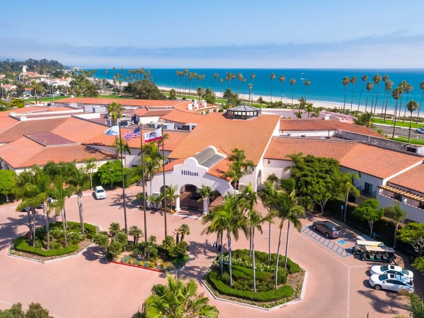Aerial view of a Hilton hotel with a red-tile roof, palm trees, and a beach with ocean view in the background.