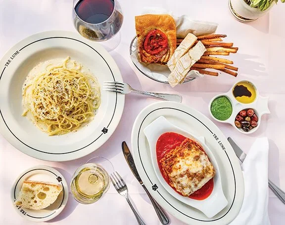 A table with a plate of spaghetti in creamy sauce, glass of red wine, a slice of bread, a plate of chicken Parmesan with tomato sauce and melted cheese, a glass of white wine, and a small bowl of mixed olives, herbs, and dipping sauces.