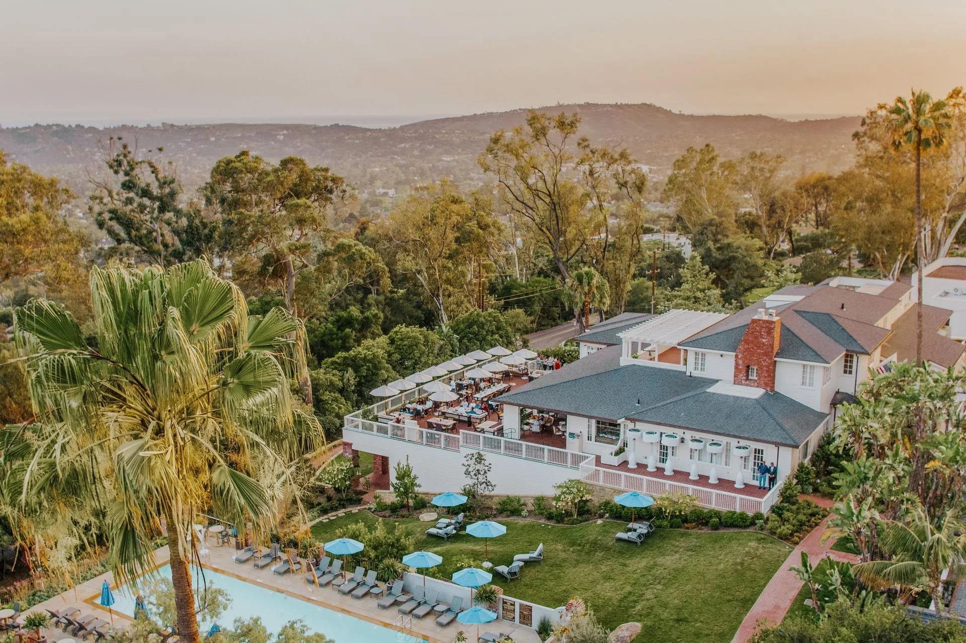 Aerial view of a large house with a pool, outdoor seating area, and lush greenery on a hillside with trees and mountains in the background.