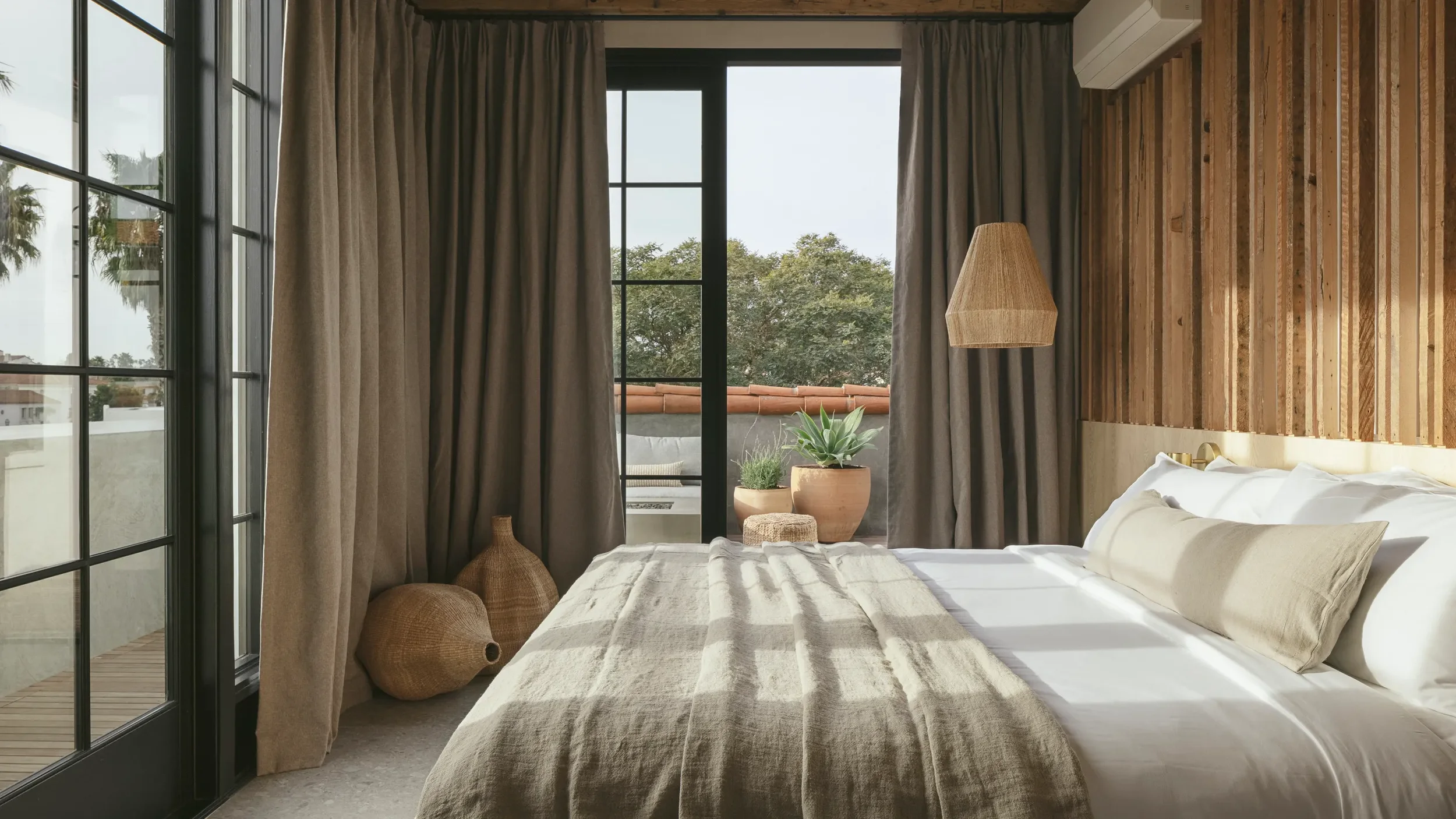 Sunlit bedroom with bed, beige linens, pillows, wooden accent wall, large windows with gray curtains, potted plants outside, wicker vases, and a hanging rattan light