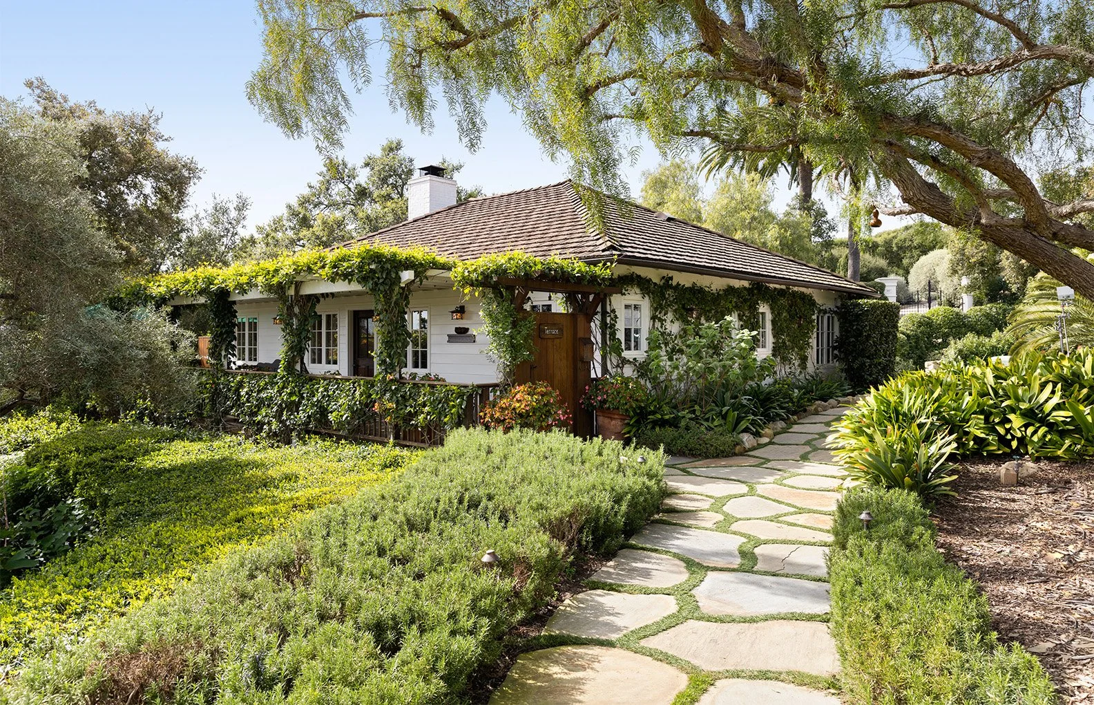 A charming white cottage surrounded by lush green plants and a stone pathway leading to the front door, with trees and clear blue sky in the background.