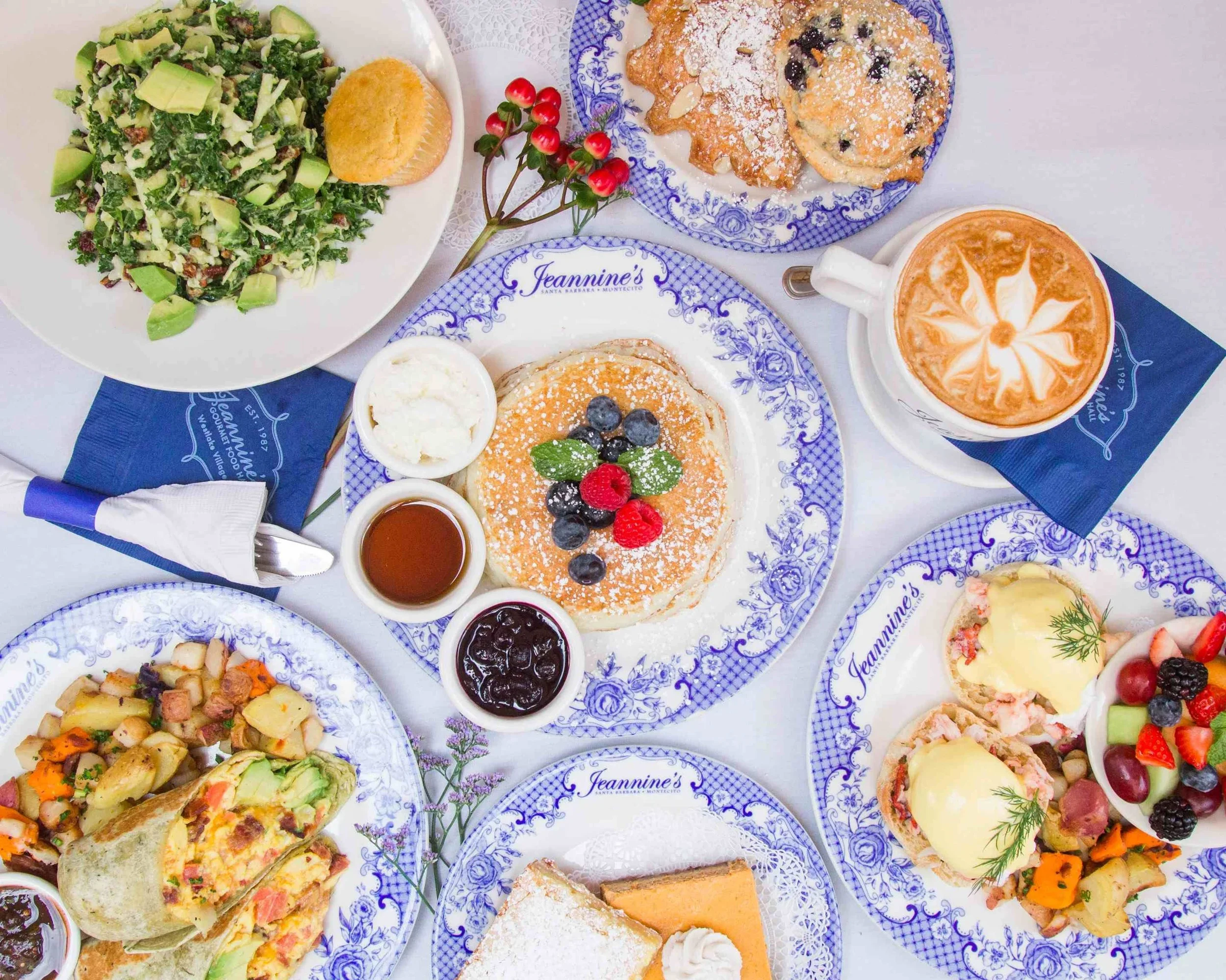 A table with various breakfast foods, including a salad, pancakes with berries, a cup of coffee, fruit salad, muffins, and a baked dish, all on blue and white patterned plates and cups.
