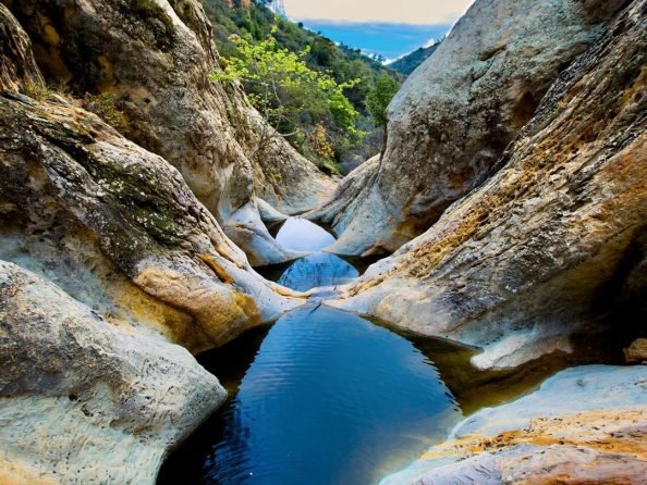 A narrow rocky canyon with a small water stream flowing through it, surrounded by large boulders and green trees in the background.
