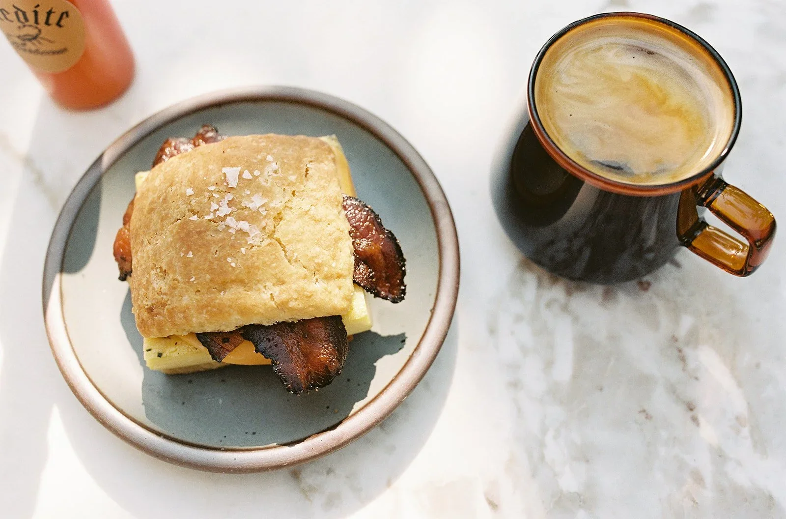 Bacon, cheese, and egg sandwich with a biscuit on top, served on a small bowl, with a dark mug of coffee or tea and a bottle of hot sauce in the background, all on a marble surface.