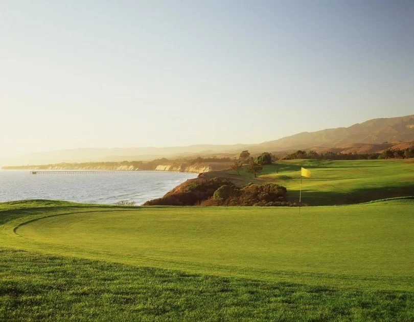 Scenic view of a golf course near a body of water with a yellow golf flag, rolling green hills, trees, and mountains in the distance.