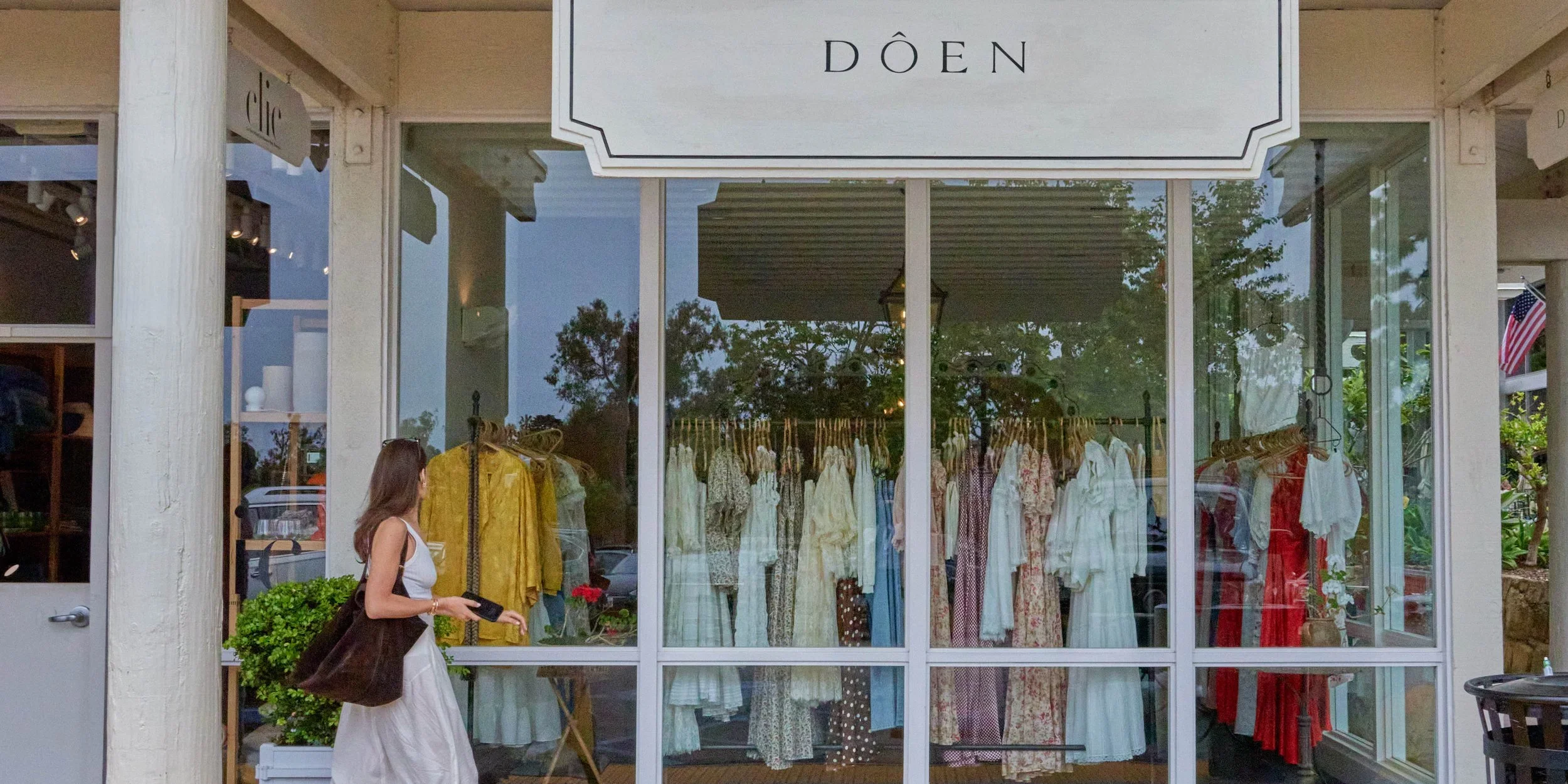 Woman in white dress walking past a clothing store window displaying dresses on racks inside.