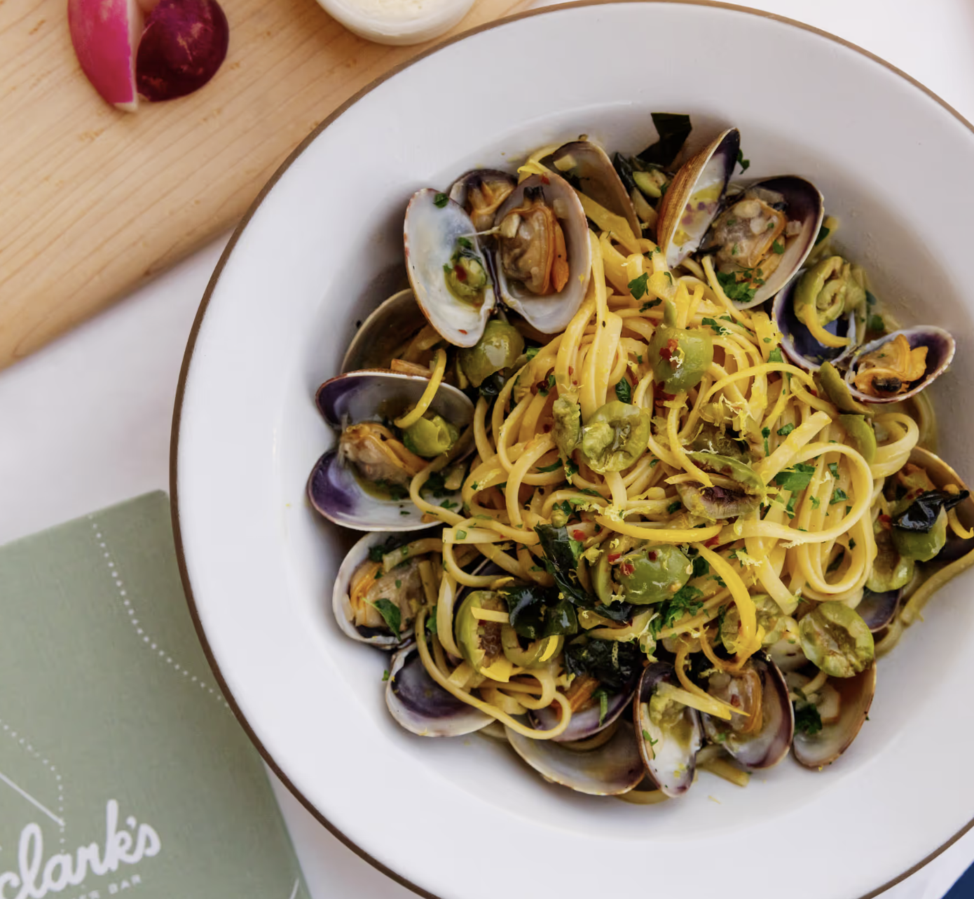 Bowl of pasta with clams, green olives, herbs, and red pepper flakes.