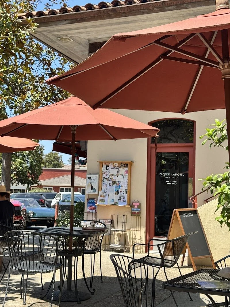 Outdoor patio with tables, chairs, and umbrellas in front of a cafe or restaurant, surrounded by greenery and trees.