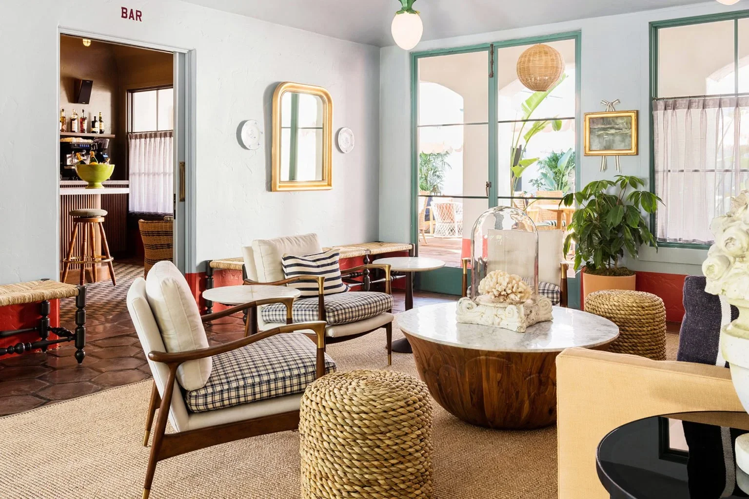 A cozy living room with mid-century modern furniture, light-colored walls, and large windows with views of a patio and greenery outside.