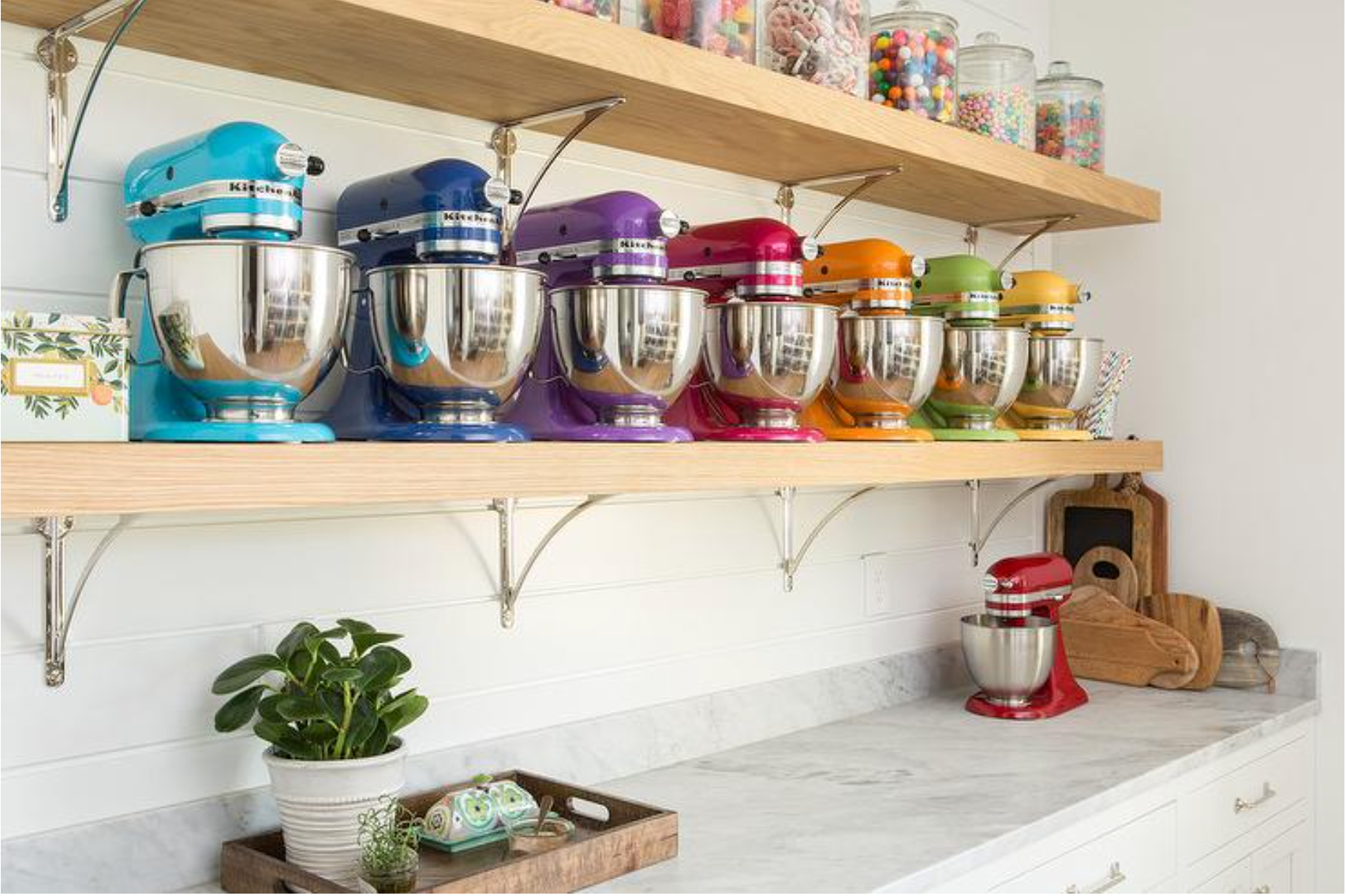 Colorful stand mixer collection on open shelves in a bright kitchen, with various baking tools and ingredients, a potted plant, and a marble countertop.