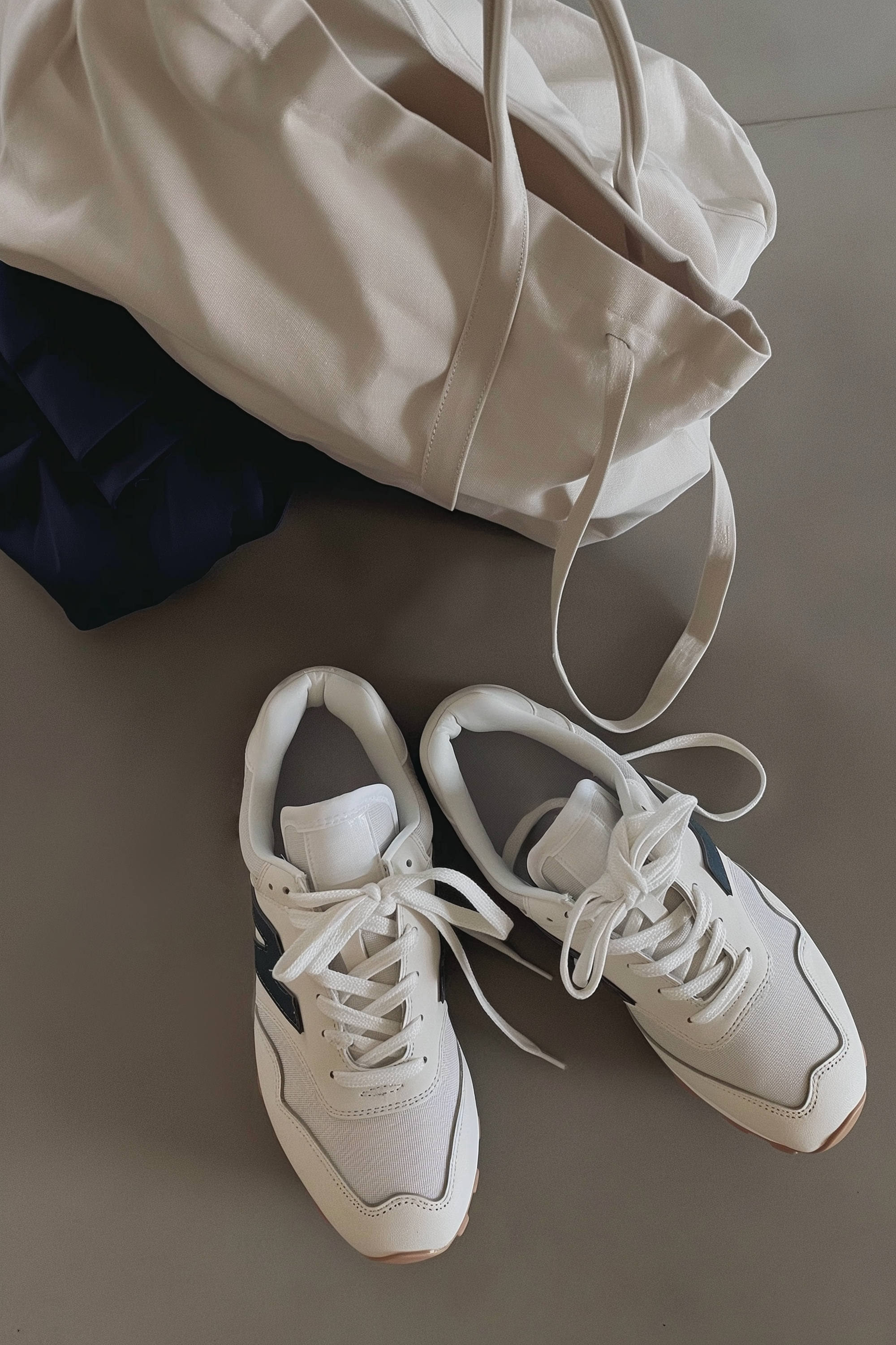 Pair of white sneakers, a white tote bag, and folded dark clothing on a gray surface.