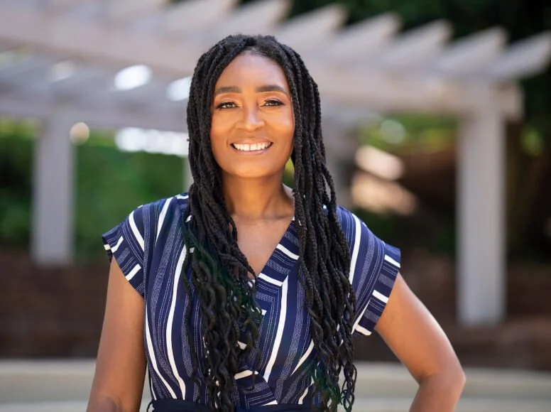 A woman with long braided hair smiling outdoors, wearing a blue striped dress.